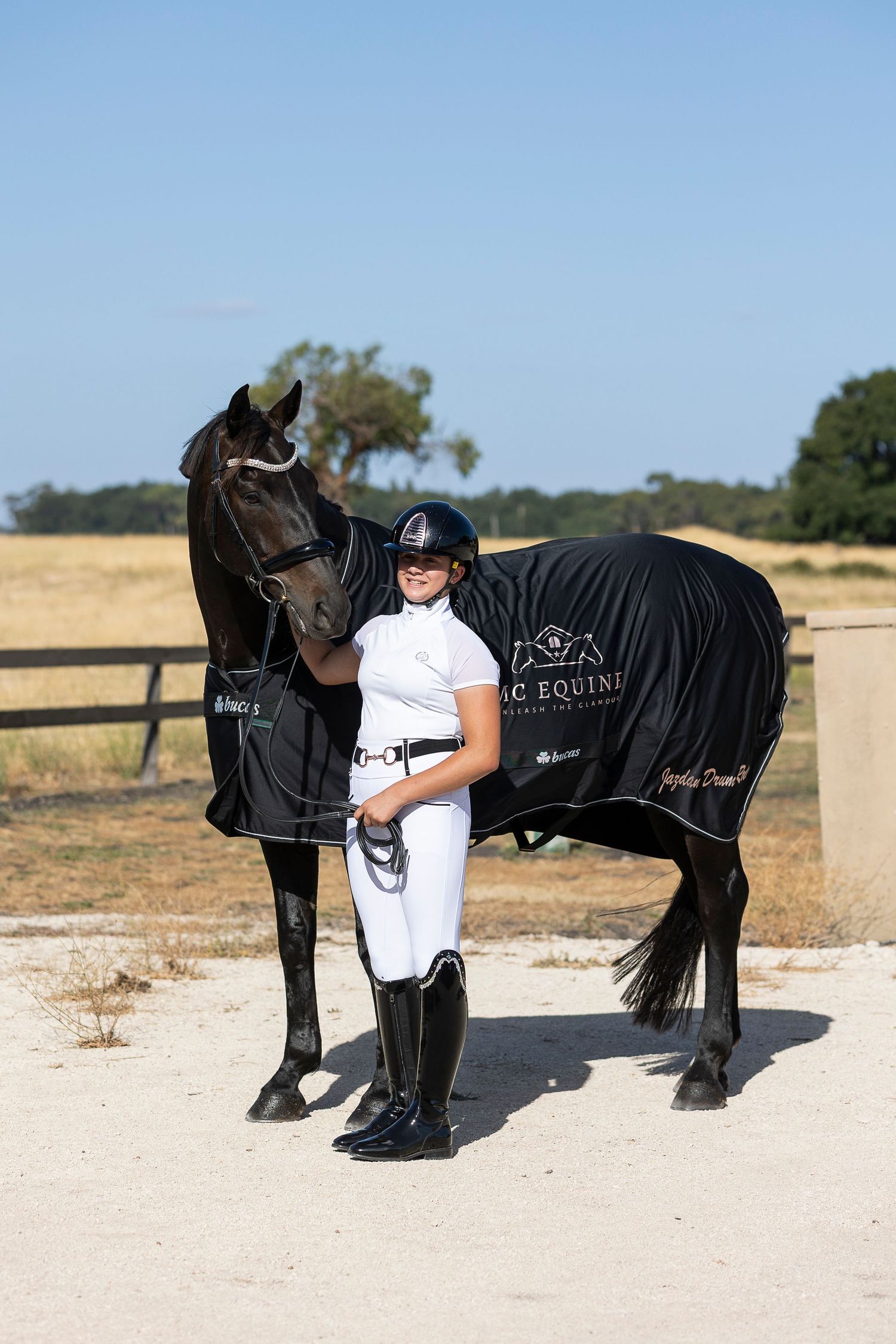 Equestrian in white competition attire standing with a black horse wearing a branded blanket.