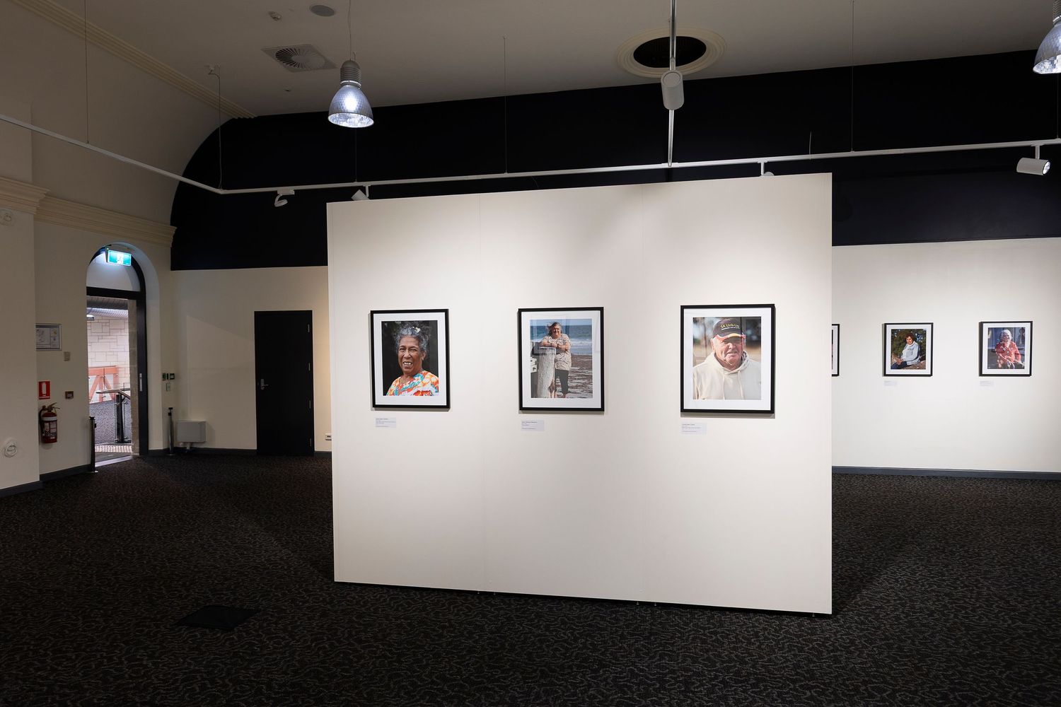 An art gallery interior with white walls displaying framed photographs in a row with track lighting overhead.