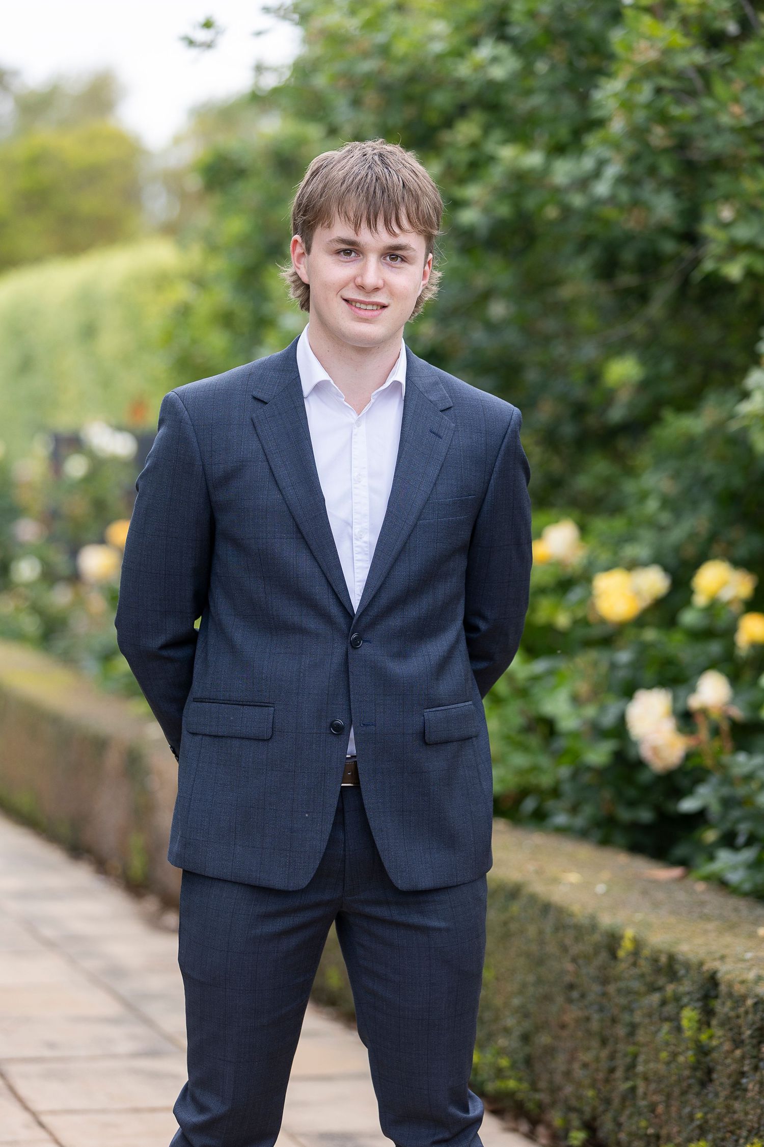 A person in a navy suit stands on a garden path near flowering bushes.