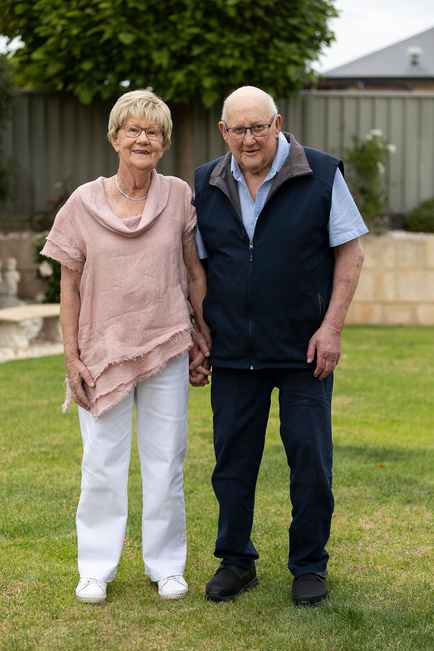 An elderly couple stands together on their lawn wearing casual attire.