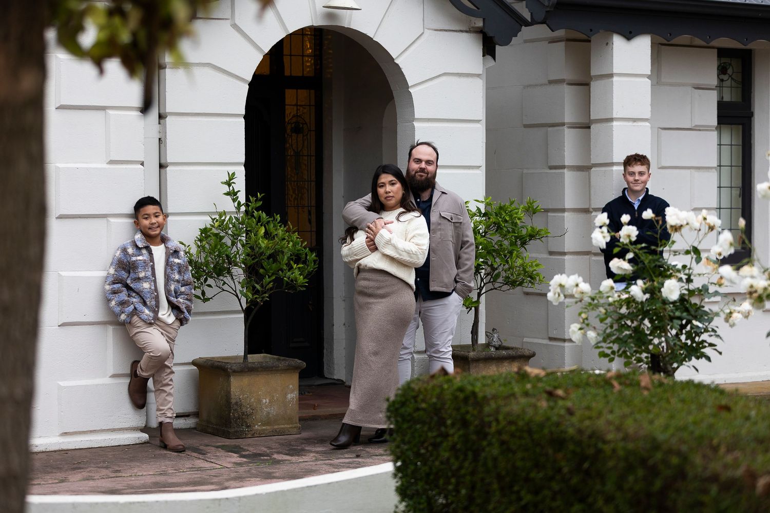Family of four poses outside elegant white manor home with lush garden, parents holding newborn baby.