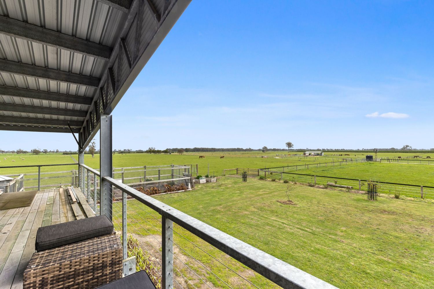 A covered balcony with wicker furniture overlooks lush green farmland and paddocks under a clear sky.