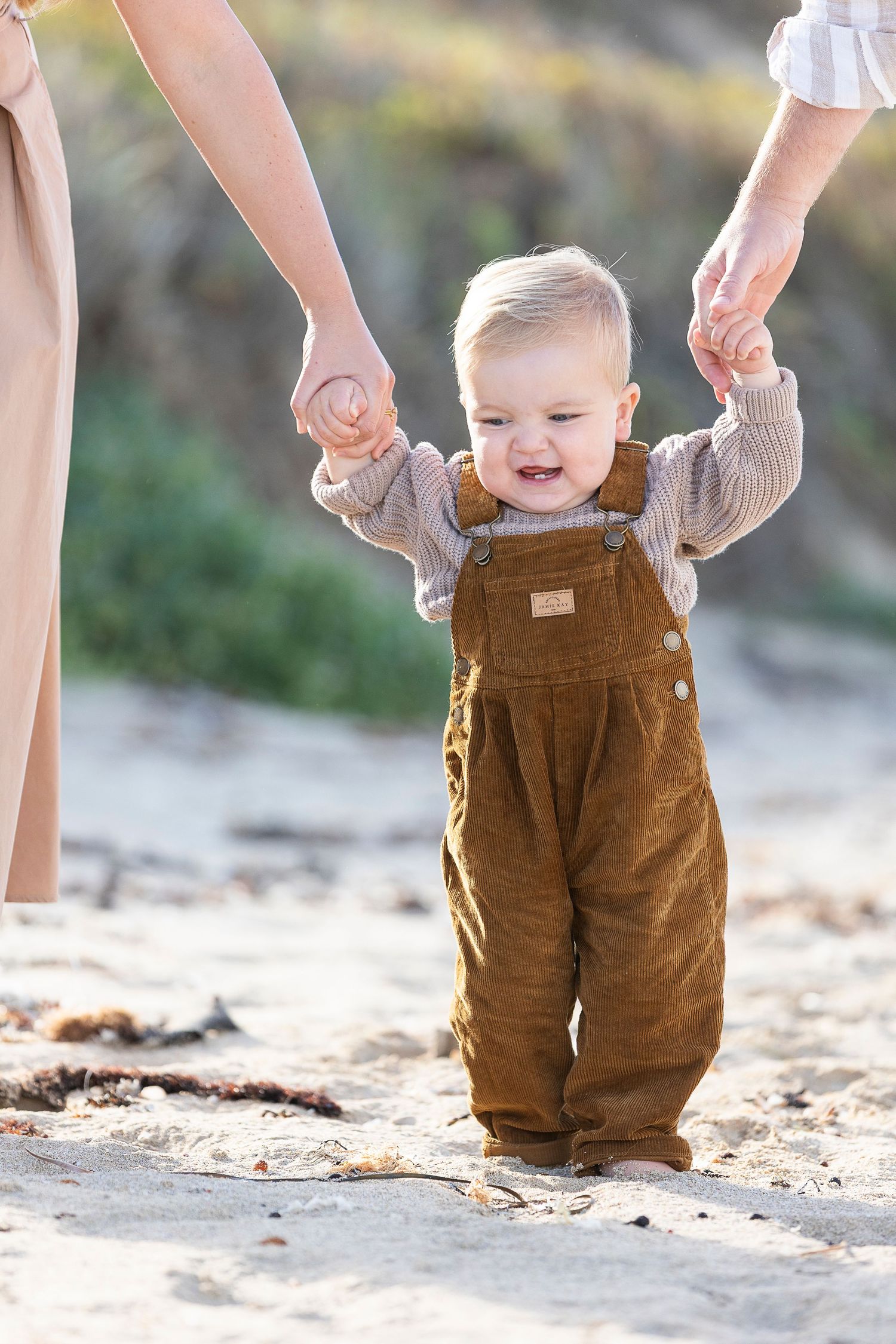 A baby in brown corduroy overalls takes first steps on the beach while holding hands.