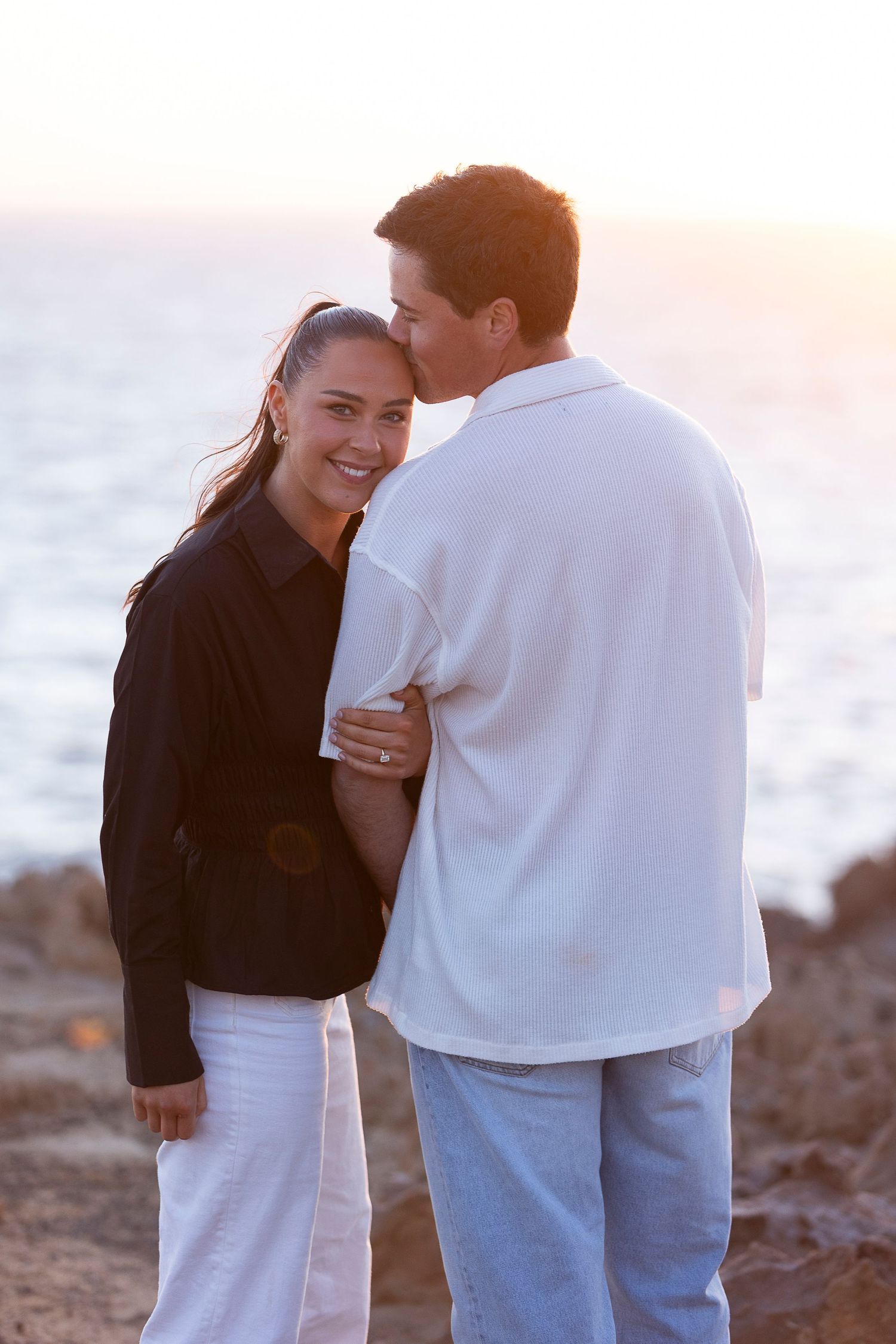 Romantic couple embraces on a rocky beach at sunset.