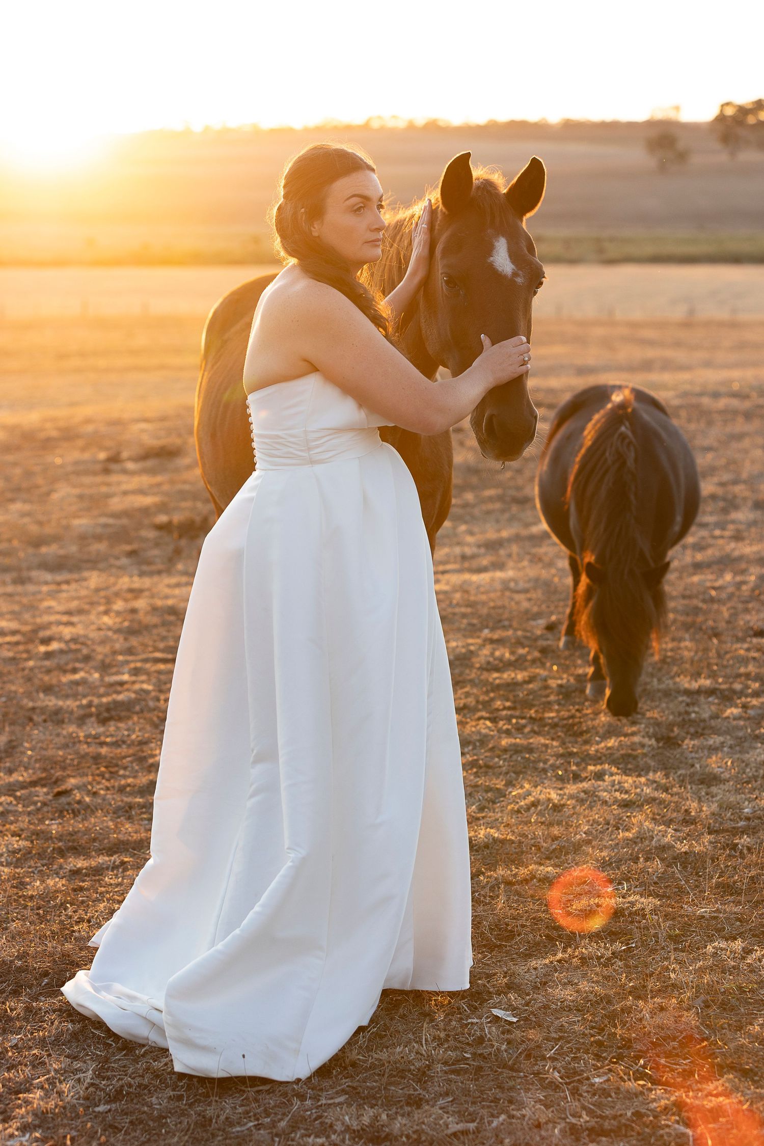 A couple in white dresses poses with a horse during golden hour.