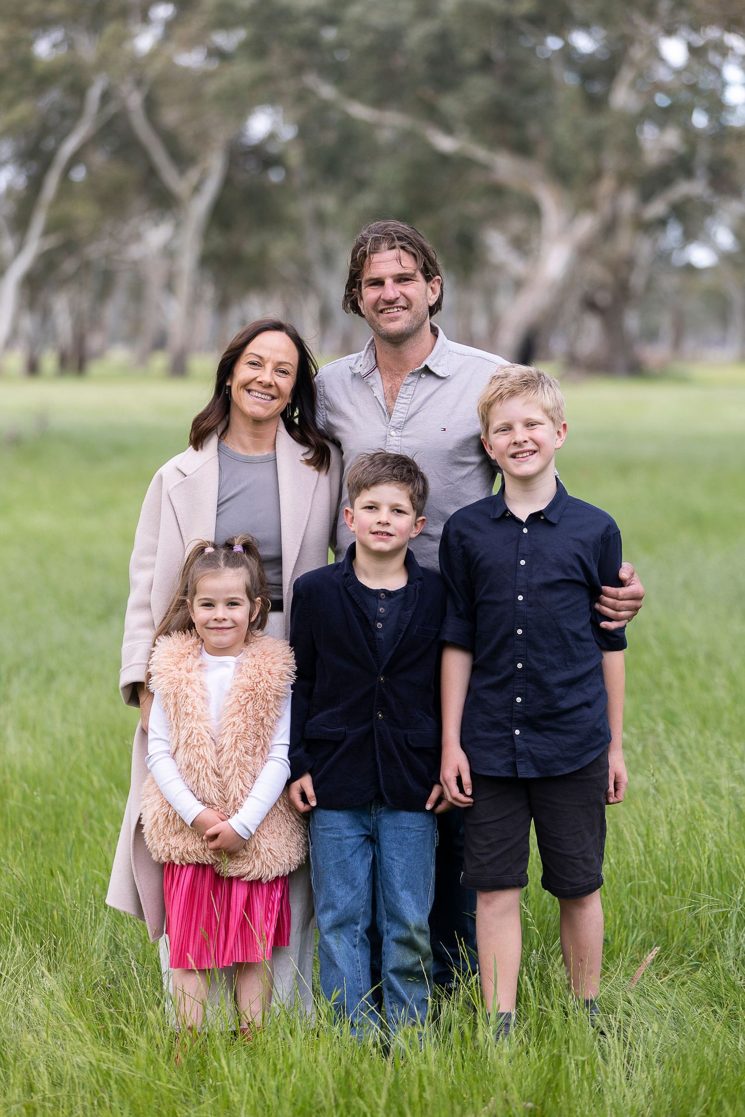 Family portrait in casual attire standing together in a grassy field with trees in the background.