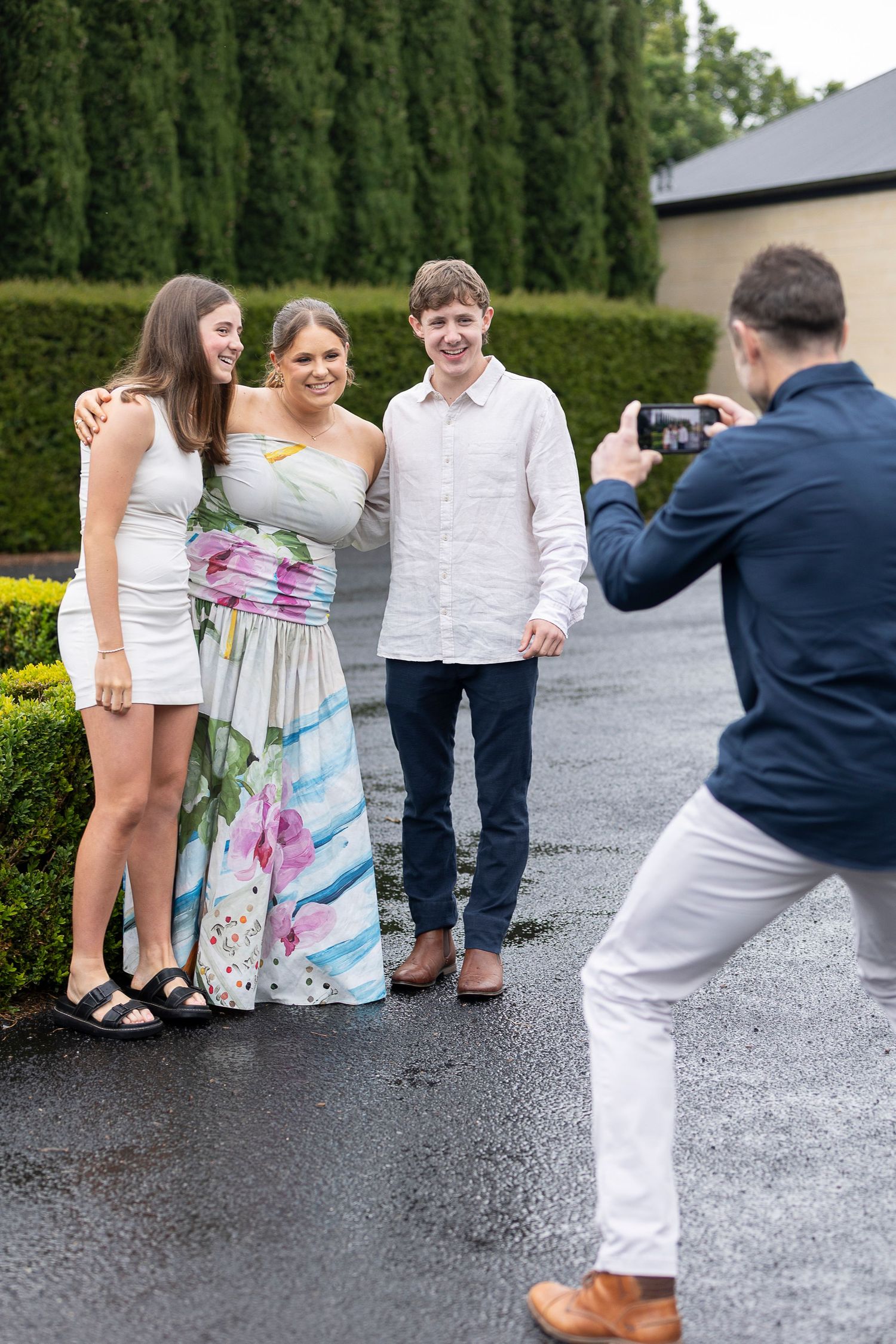People in formal attire pose playfully on wet pavement near hedges.