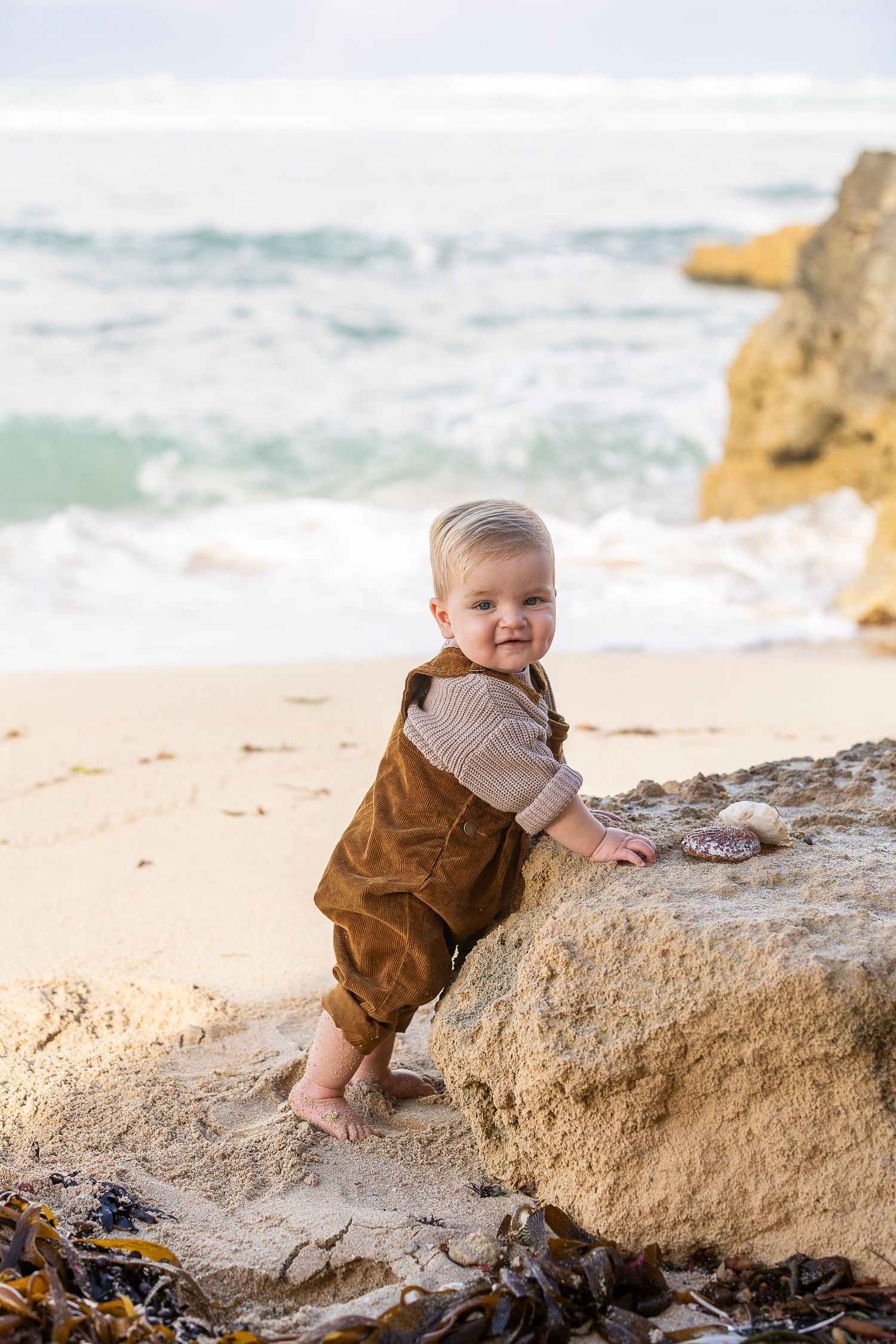 A baby in brown overalls explores a large rock formation on a sandy beach.