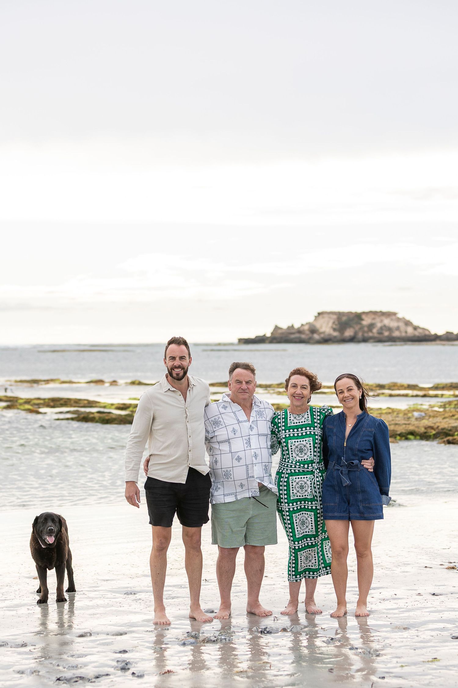 A group poses with their black dog on a beach with an island visible in the background.