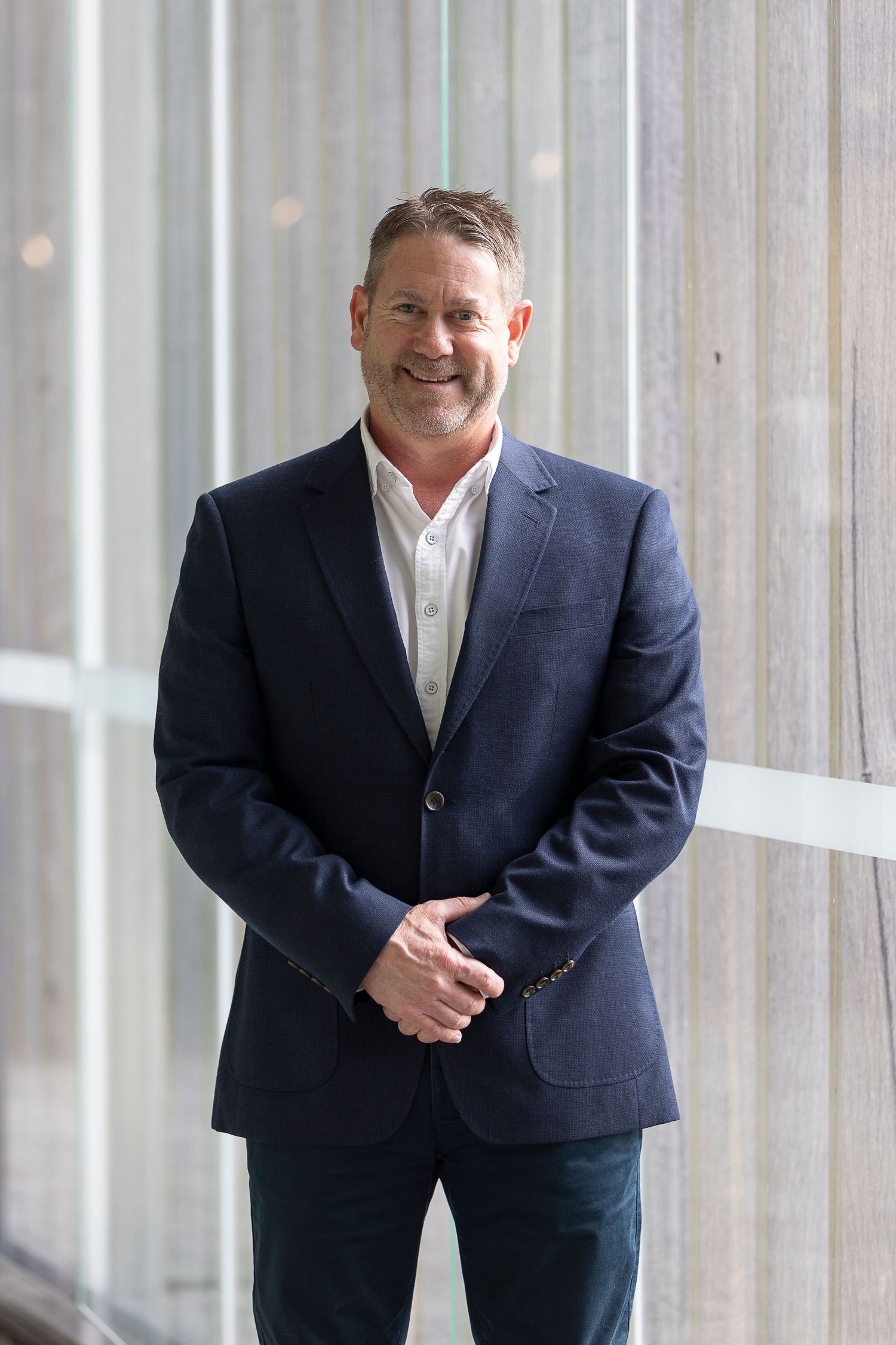 Smiling middle-aged man in navy blazer and white shirt standing confidently indoors near large windows.