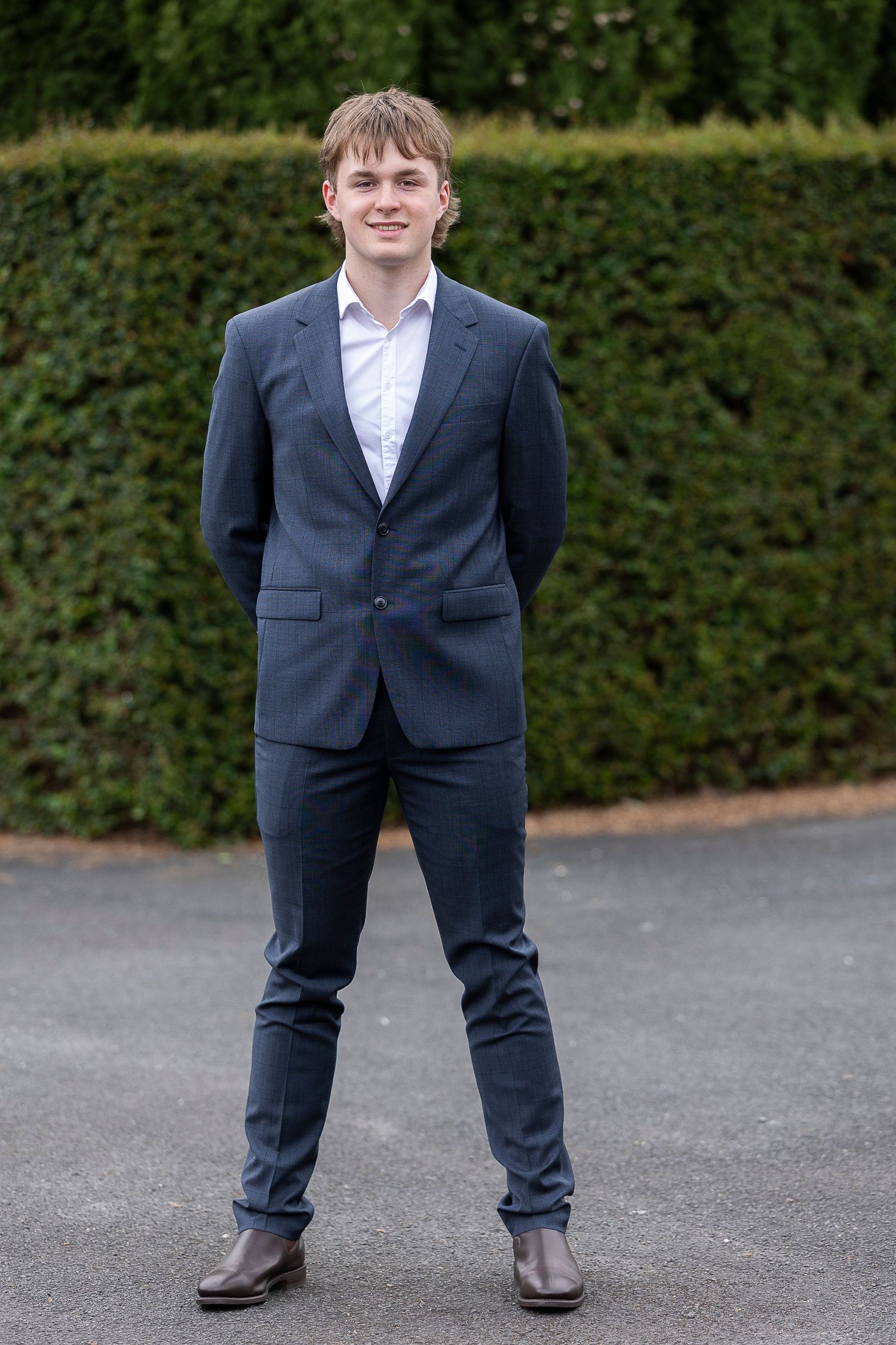 A young man in a navy suit stands on pavement in front of a manicured hedge.