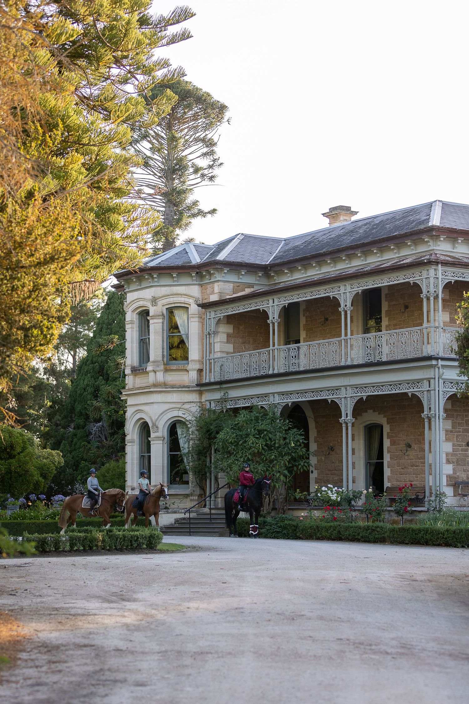 Historic two-story Victorian mansion with ornate balcony, sandstone facade, and horse riders on gravel driveway surrounded by tall trees.