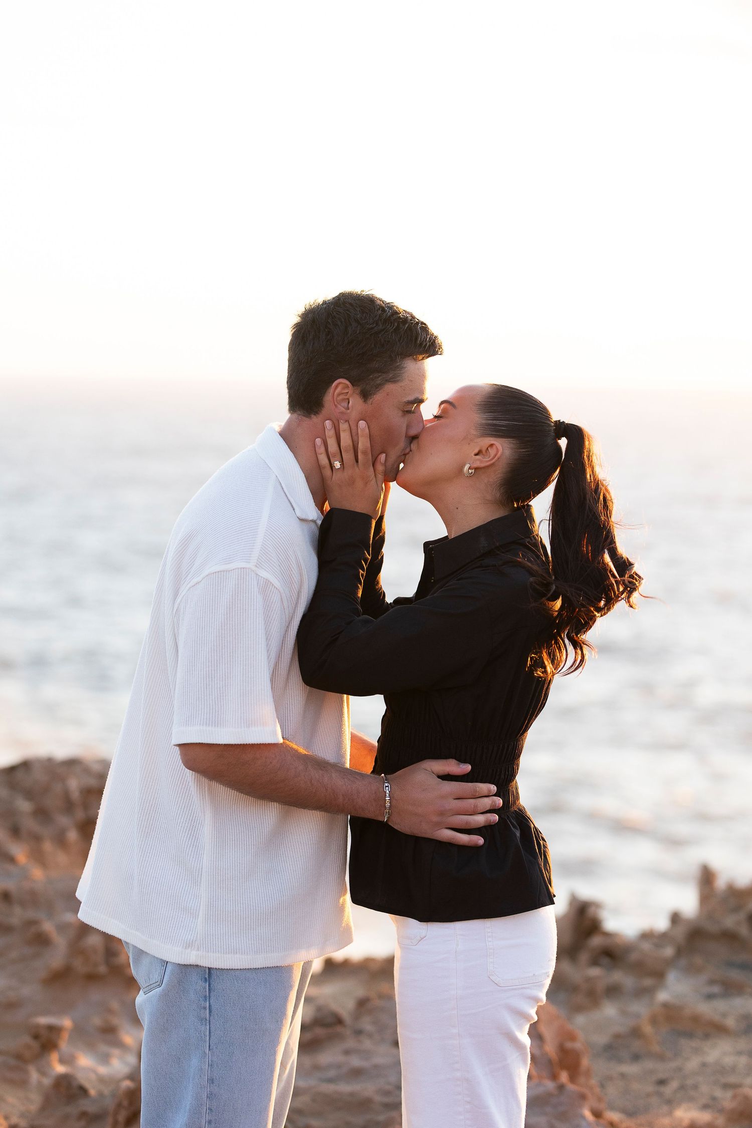 A couple shares a romantic kiss on a rocky beach at sunset.
