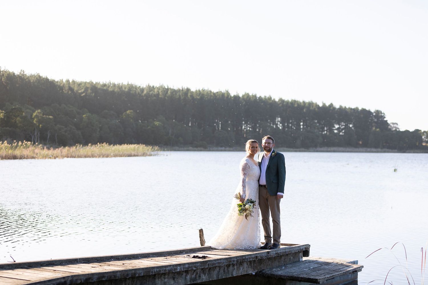 A distant view of a couple in wedding attire standing on a pier with a forested shoreline.