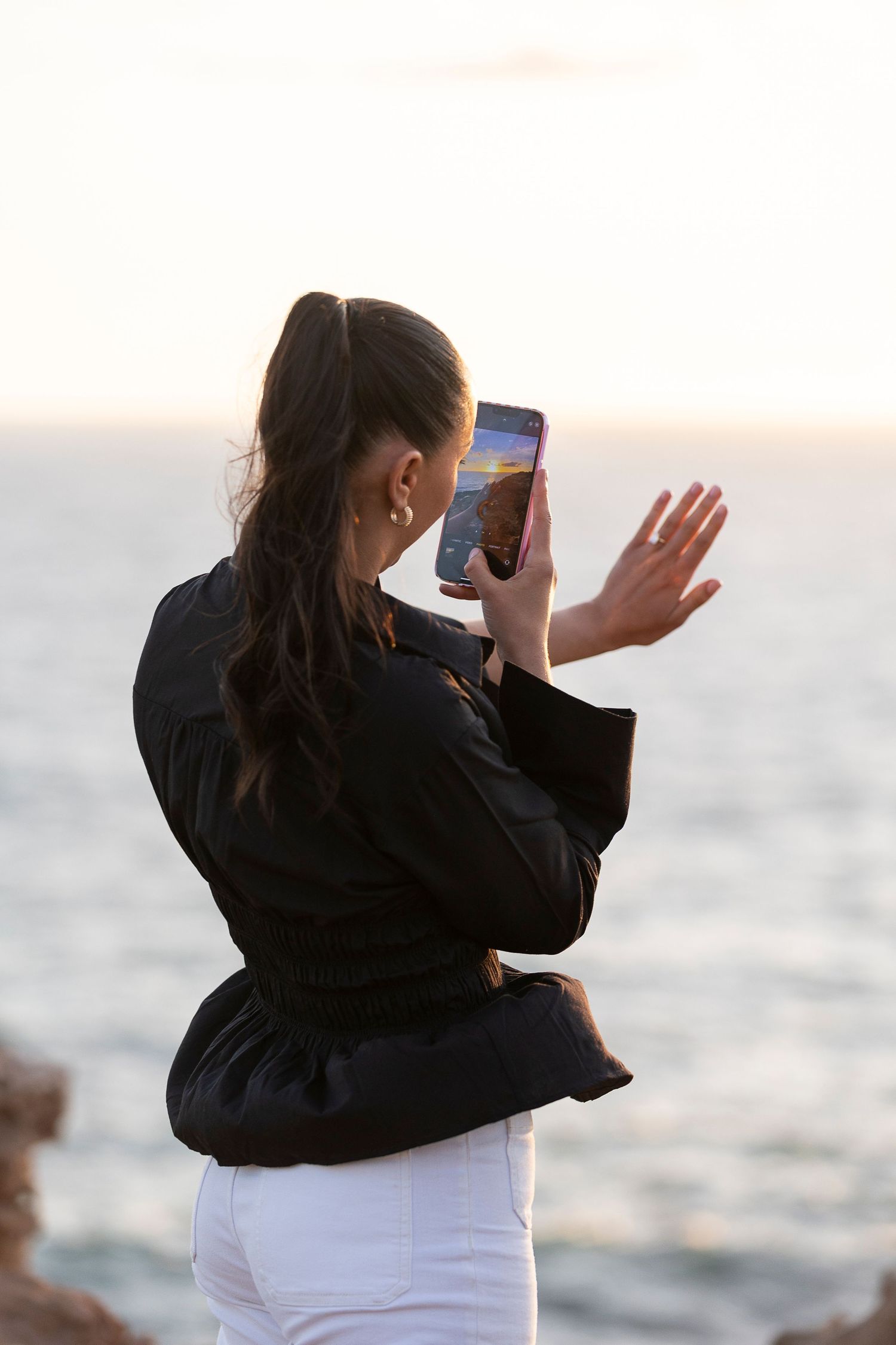 Person in black taking photos against ocean backdrop at sunset.