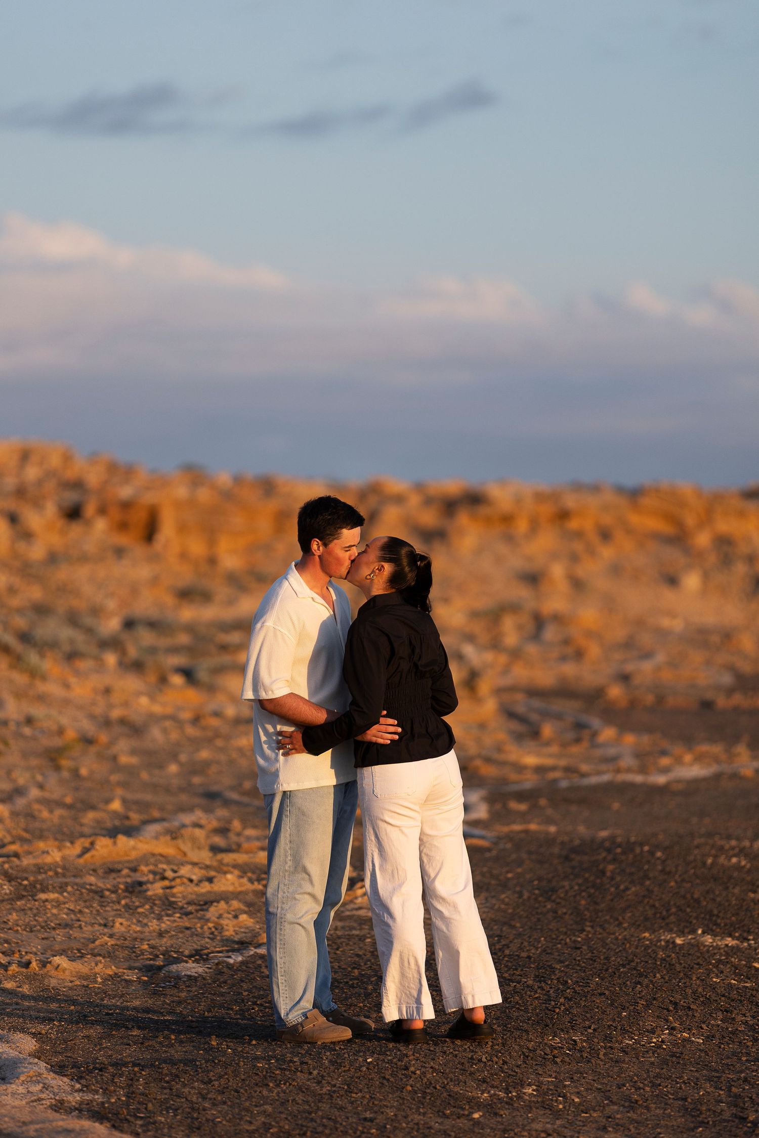 A romantic couple in white clothing embraces on a rocky beach at sunset.