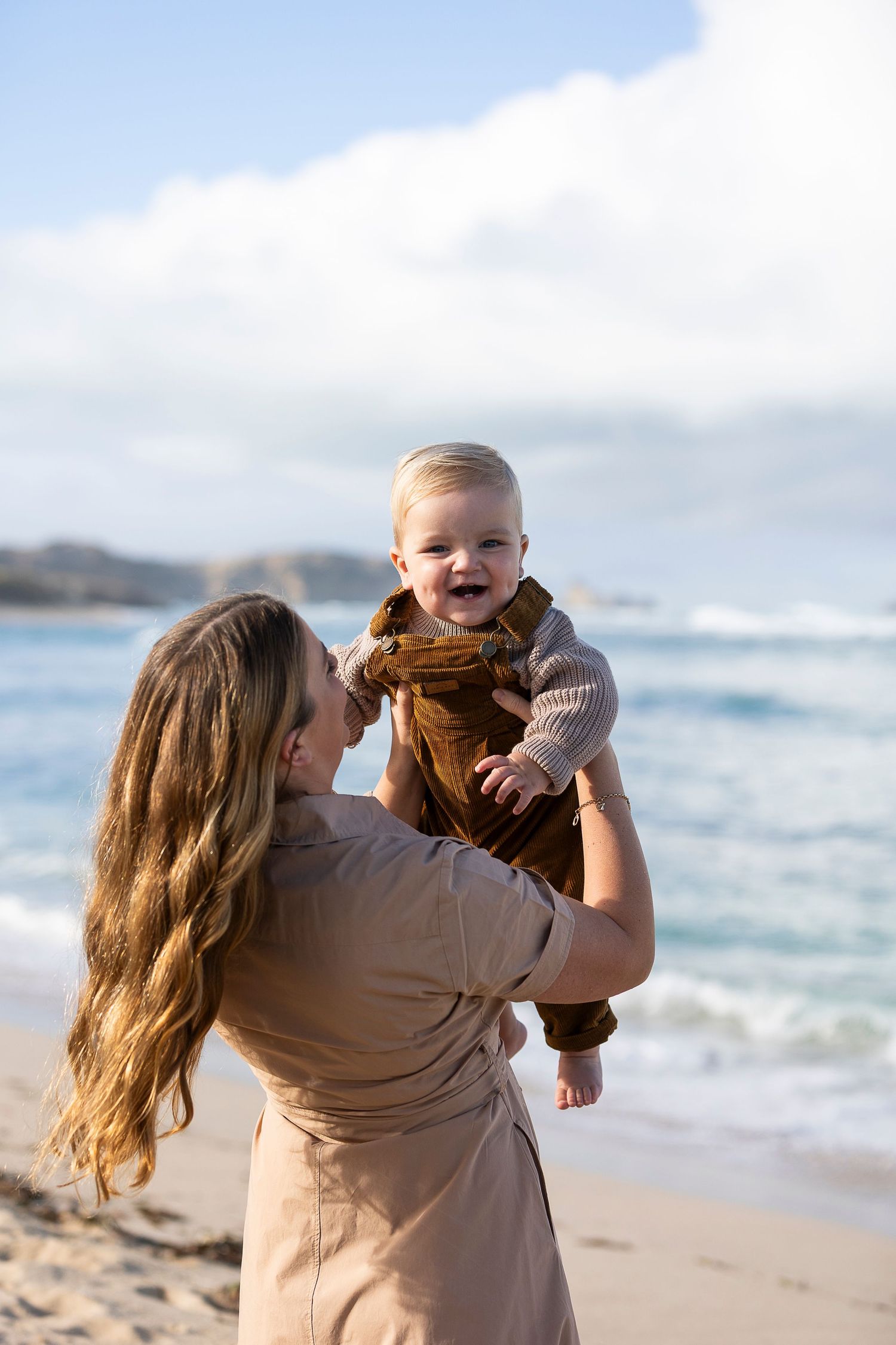 A person holds up a smiling baby wearing brown overalls at the beach.