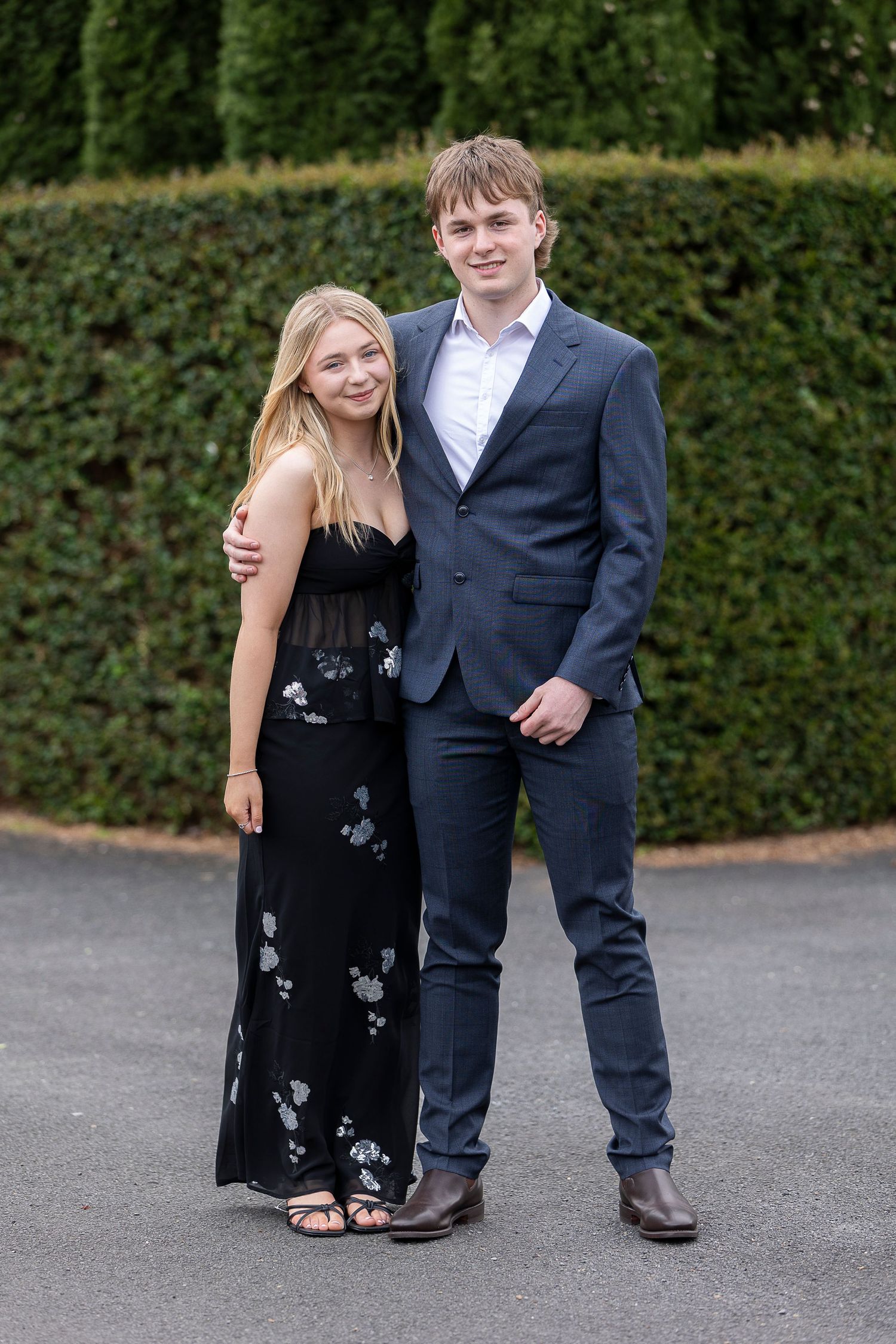 A couple in formal attire poses together in front of a green hedge.