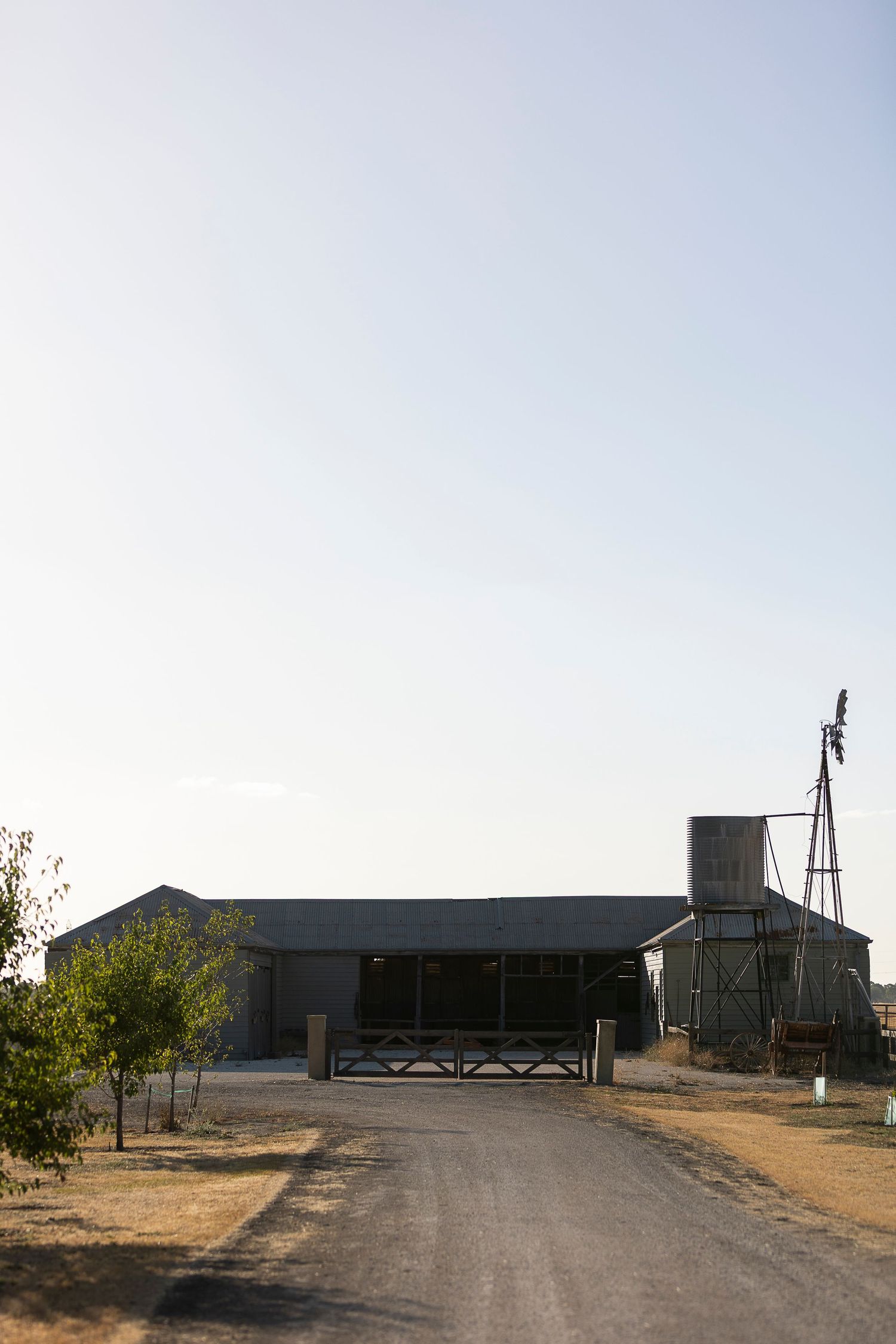 Rural farm with metal barn, windmill, silo, and gravel driveway under a clear sky.