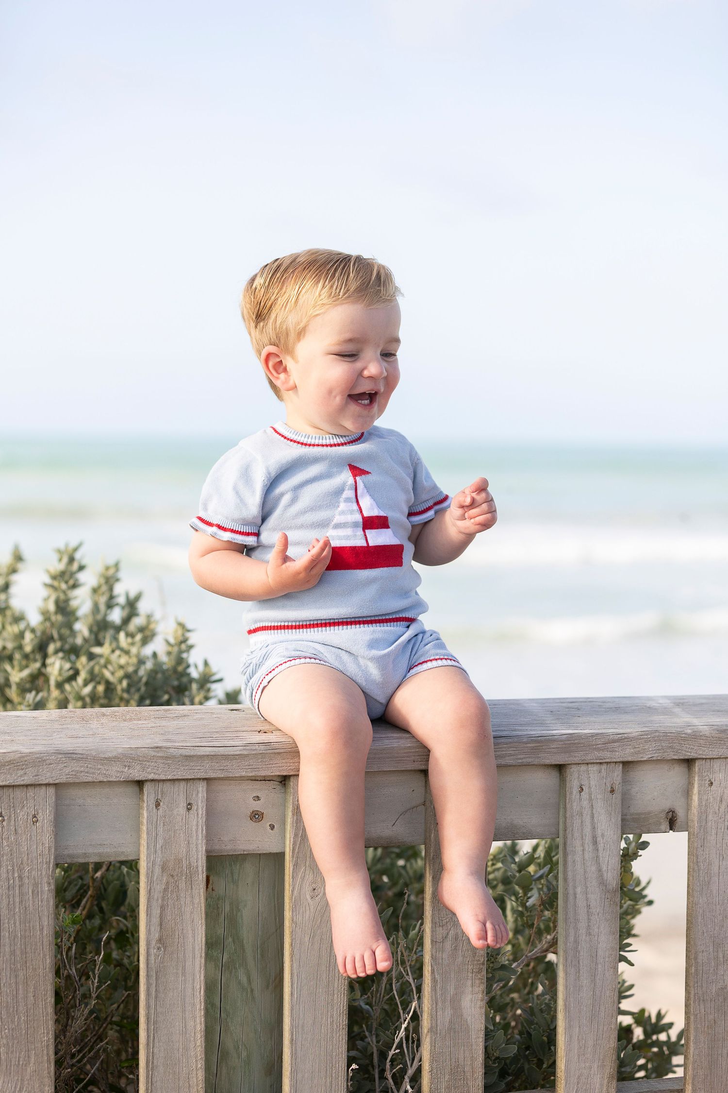 Toddler in nautical outfit sits smiling on wooden beach railing with ocean view.