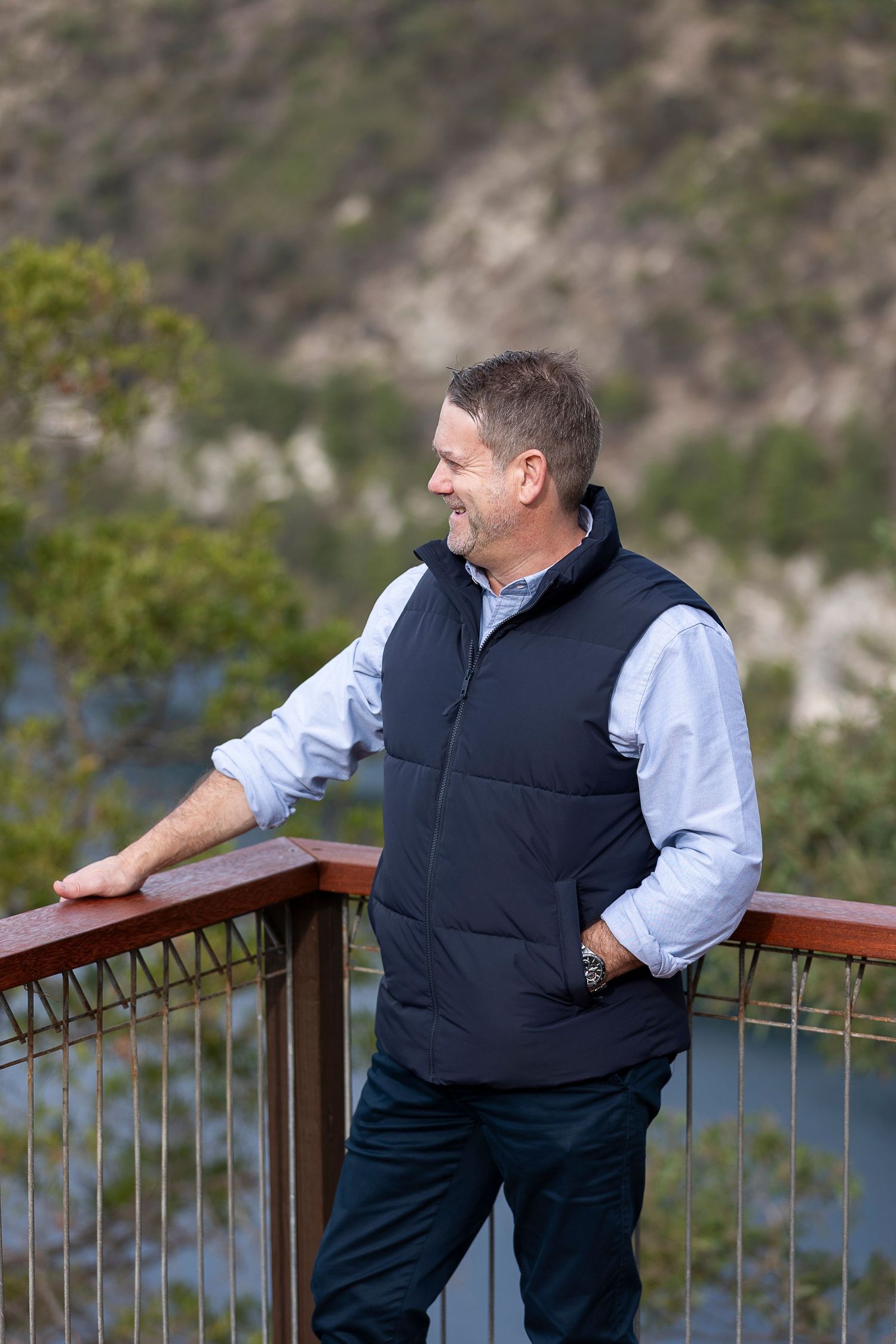 Smiling middle-aged man in navy vest and blue shirt leaning on wooden railing with scenic mountain view.