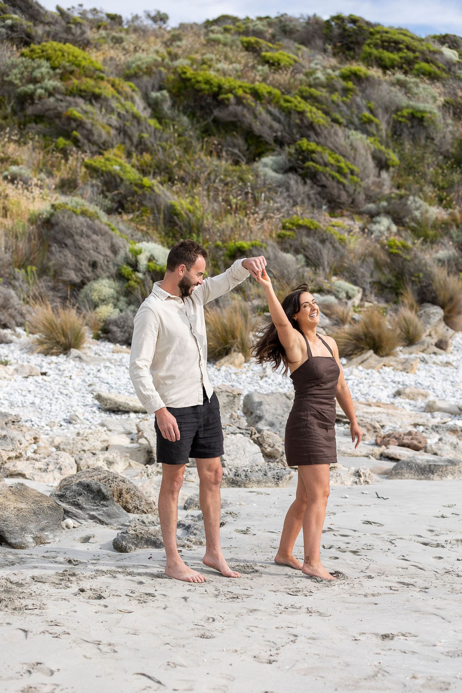 A couple dances together on a beach with rocky cliffs in the background.