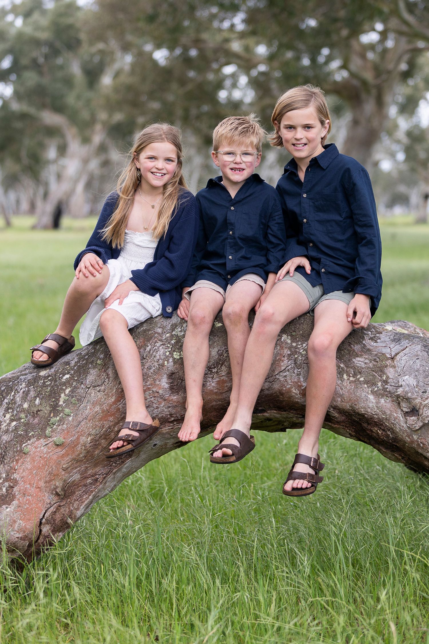 Children in matching navy outfits sitting casually on a large tree branch in a grassy field.