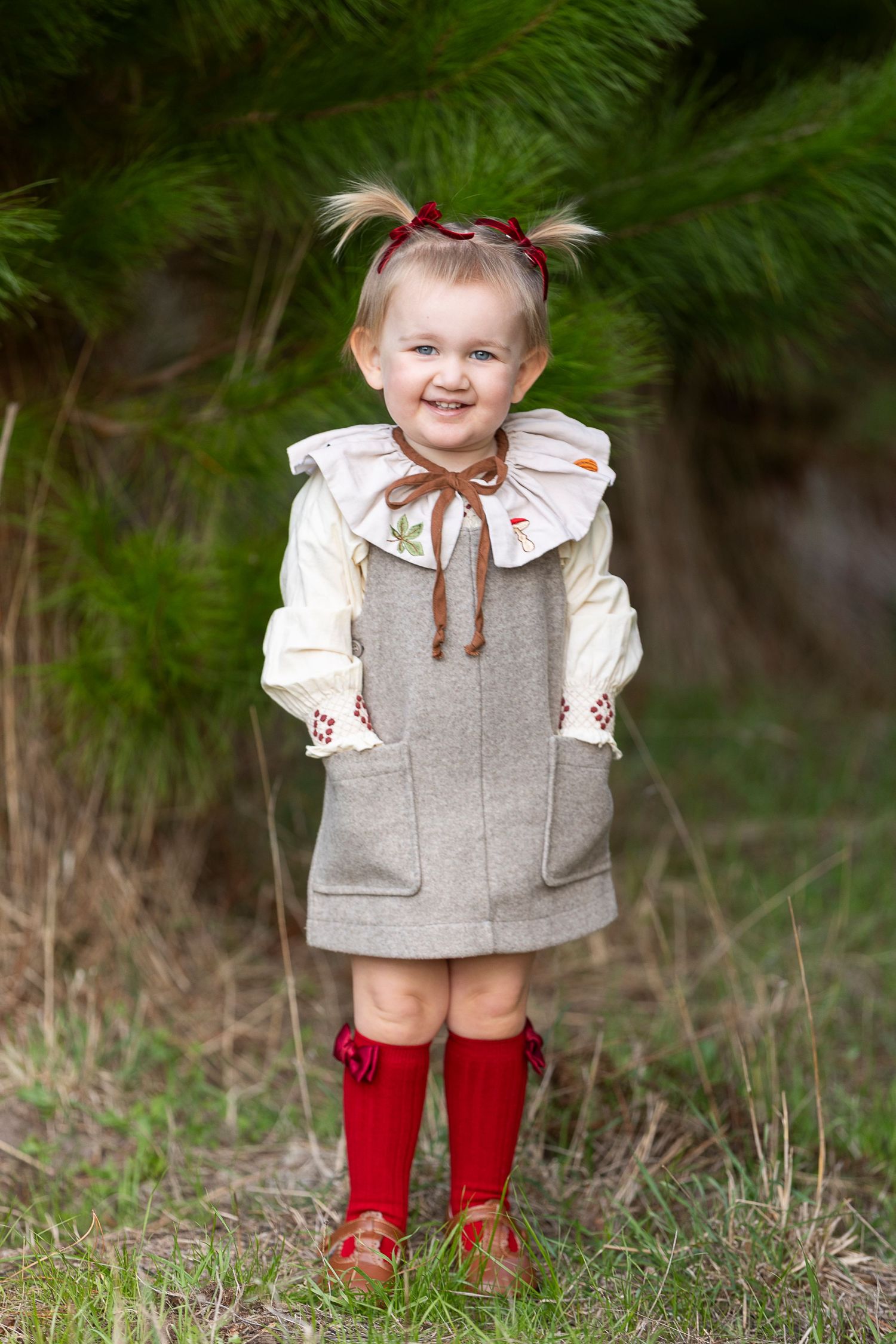 Child in vintage-style grey dress and red socks smiles in front of pine trees.