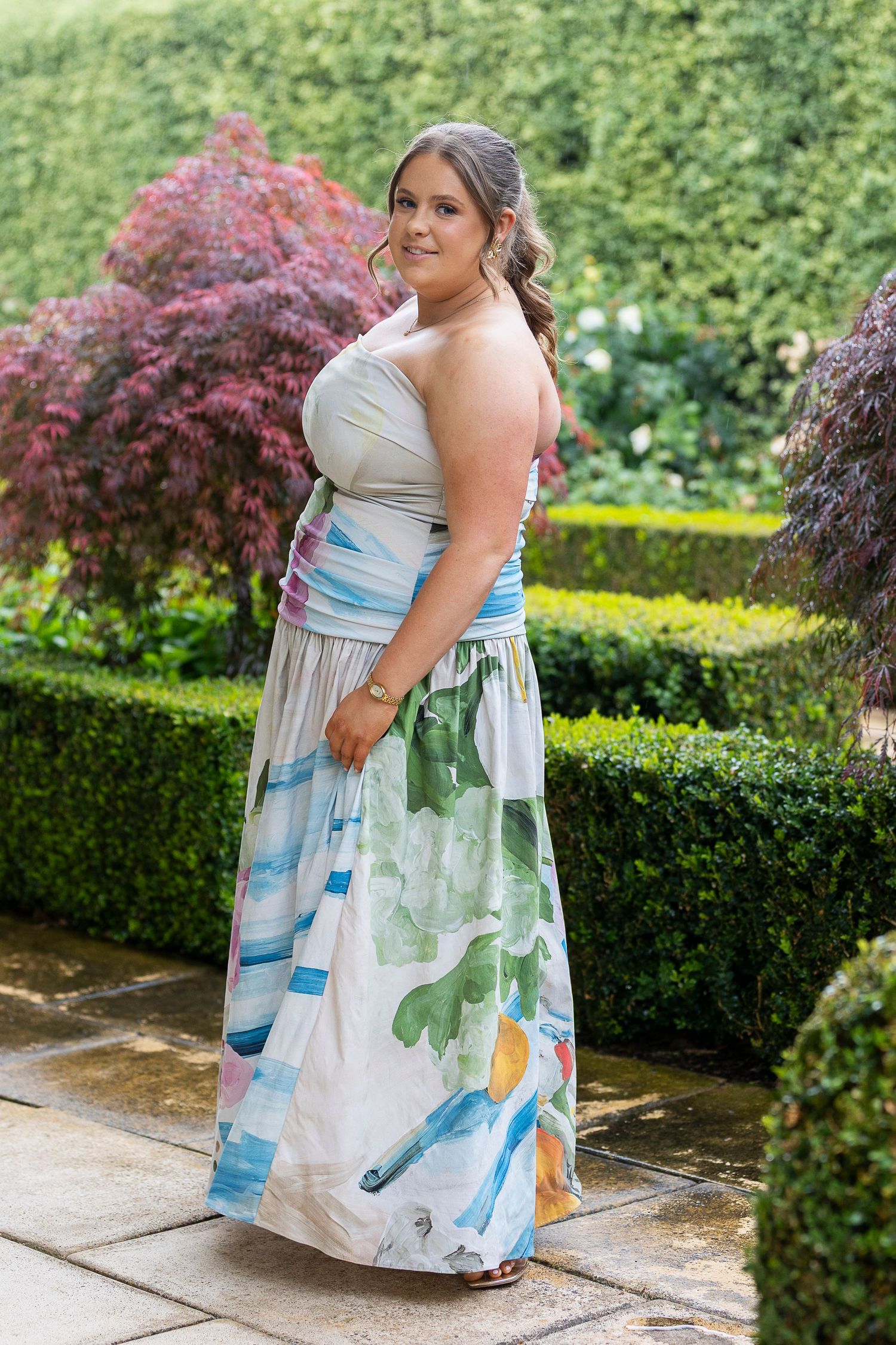 Watercolor print maxi dress styled in outdoor garden setting with hedges.