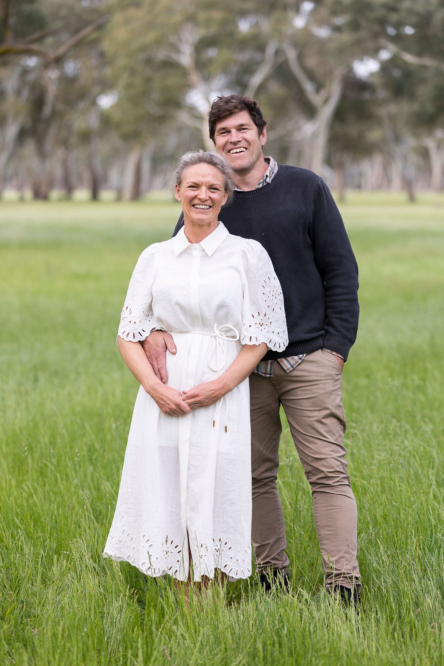 A couple poses in a grassy field wearing white and dark casual clothing.