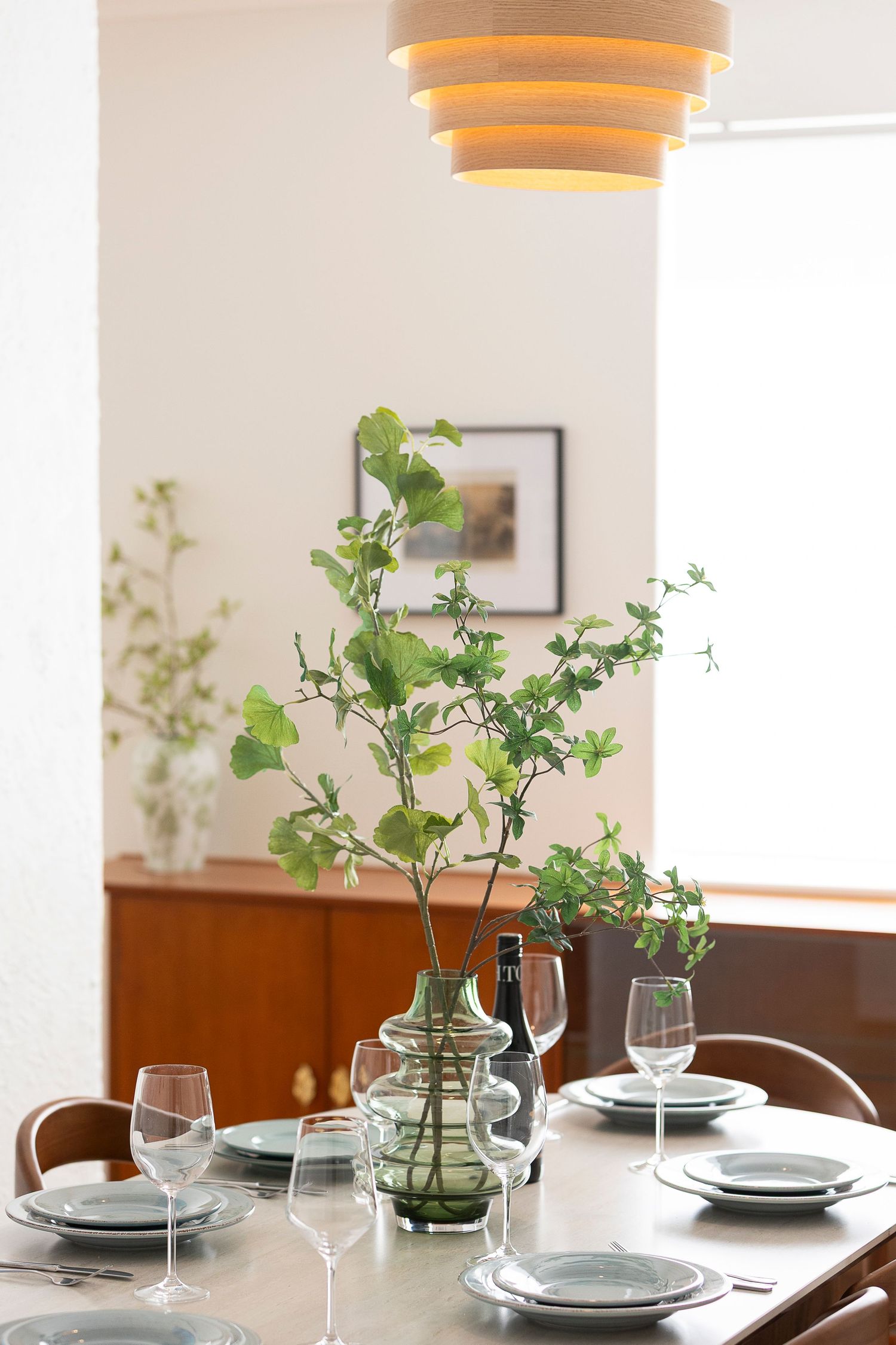 Elegant dining table set with green plant centerpiece, wine glasses, and ceramic plates under a layered pendant light.