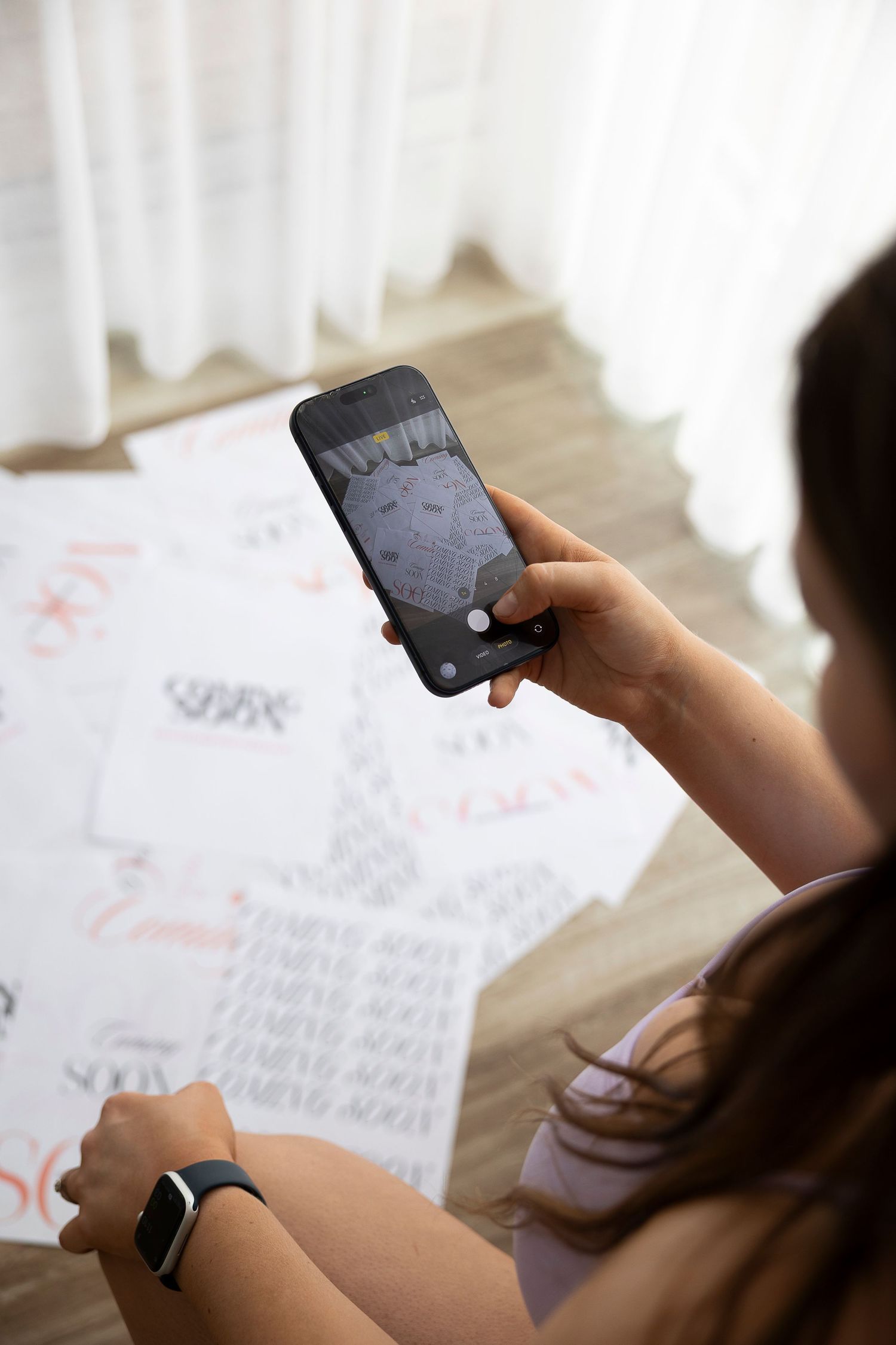Woman photographing calligraphy practice sheets on floor with smartphone while wearing a smartwatch.