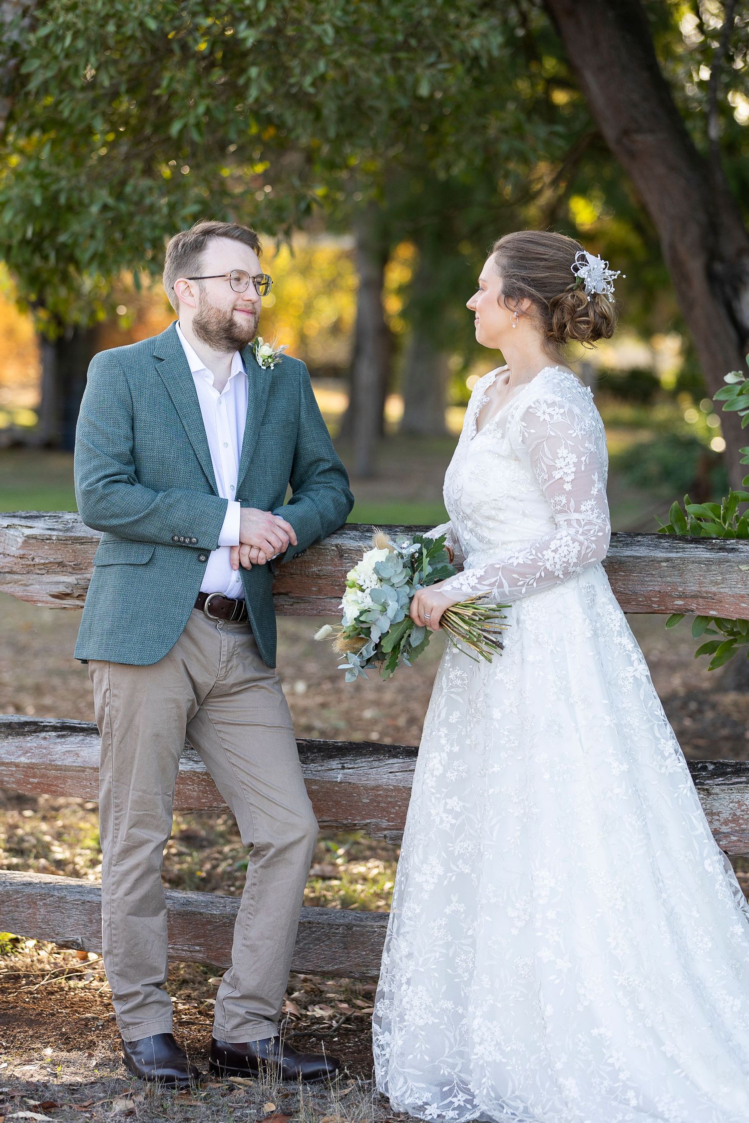 A couple in wedding attire poses together near fallen logs in an autumn garden setting.