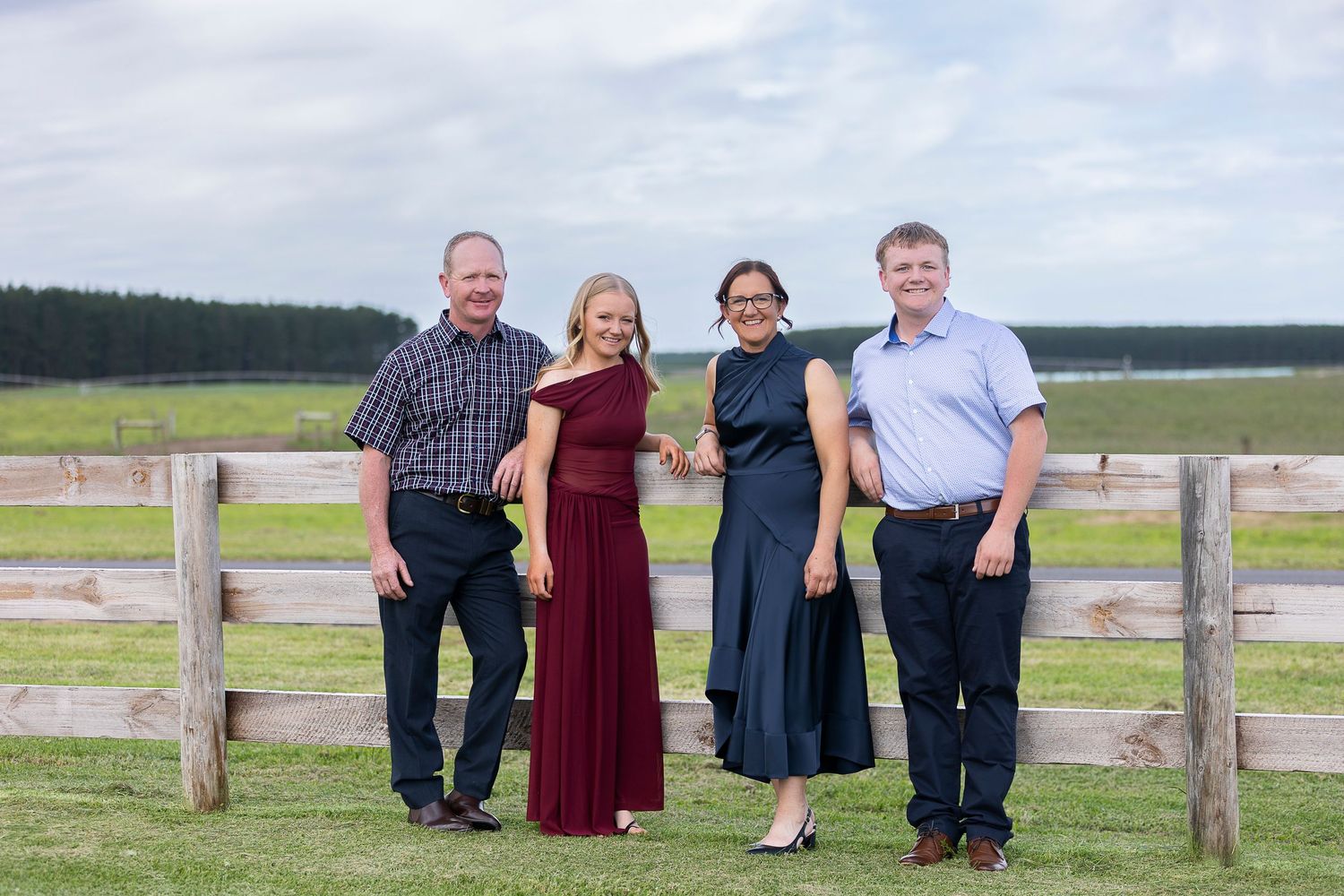 A group poses together by a wooden fence on a rural property with fields and forest in the background.