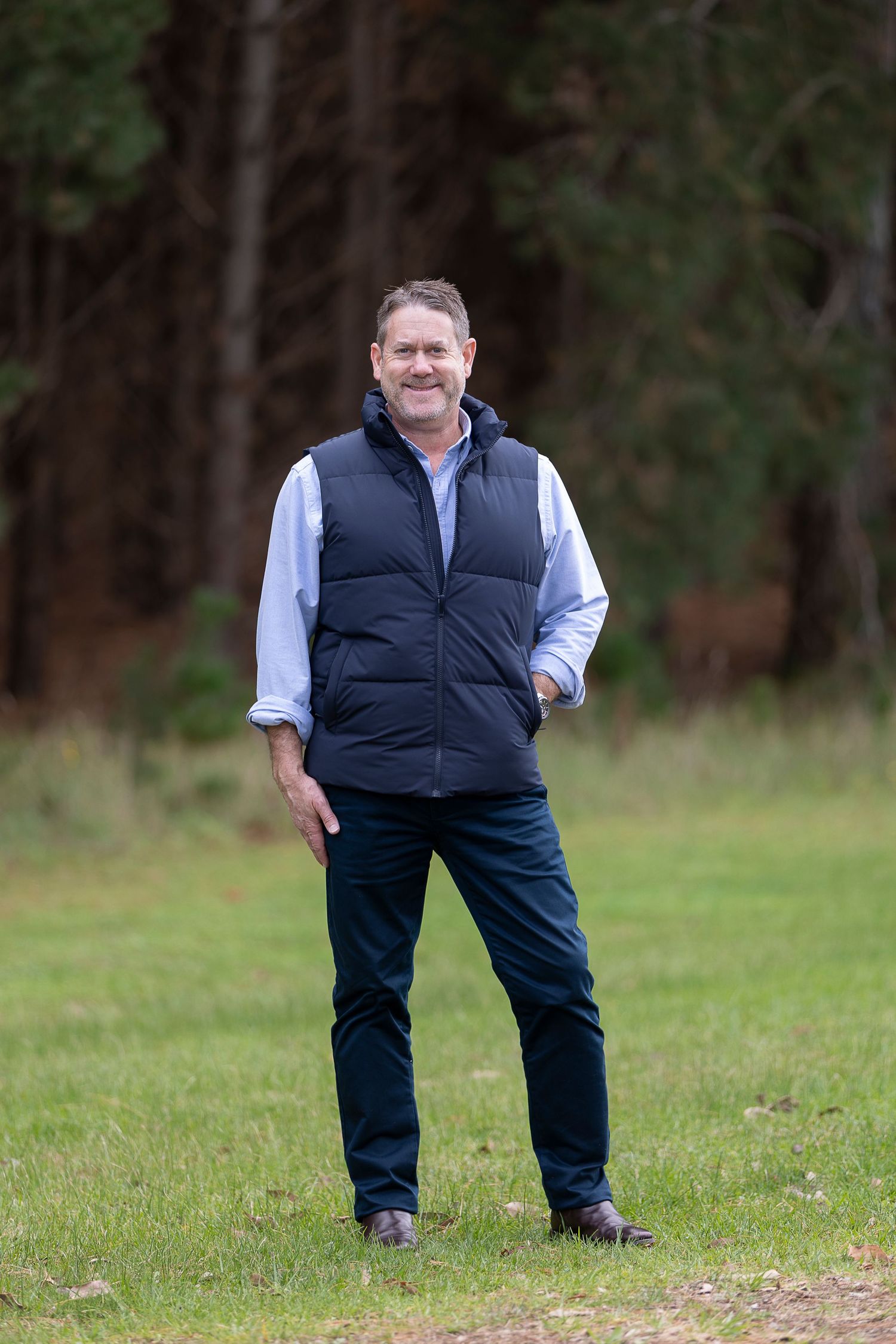 Smiling middle-aged man in navy vest and blue shirt standing on grass with forest background.