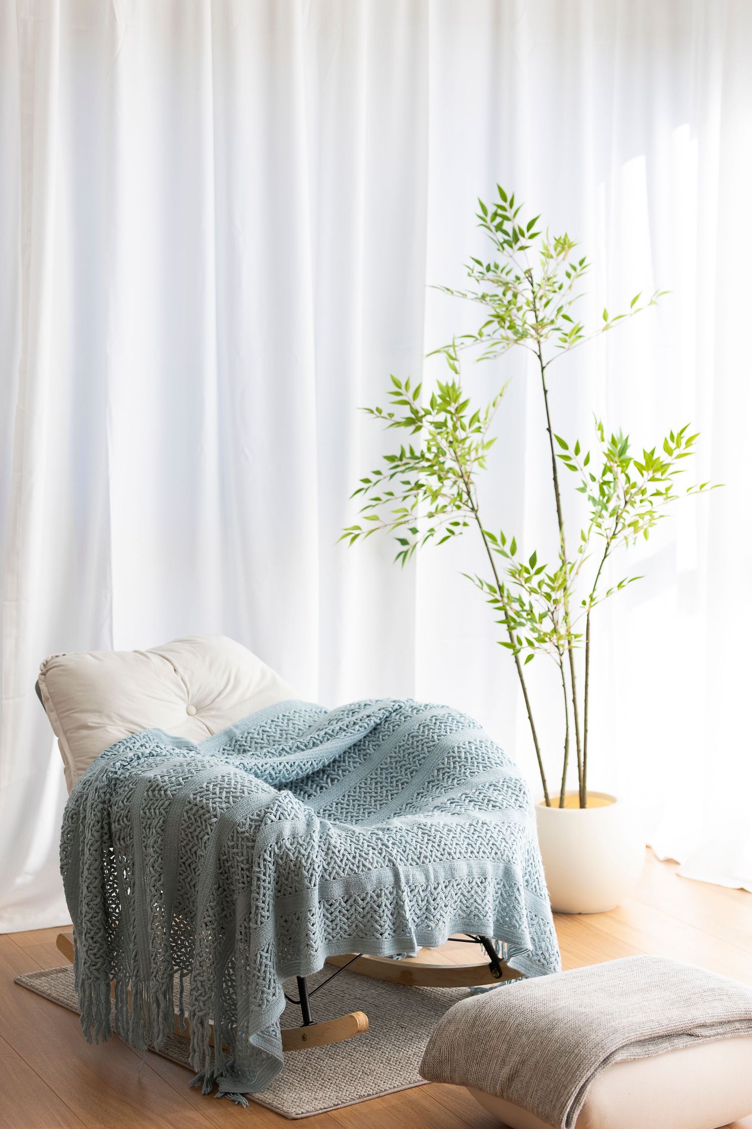 Cozy reading nook with a cream armchair, blue knit throw blanket, and tall green plant by sheer white curtains.