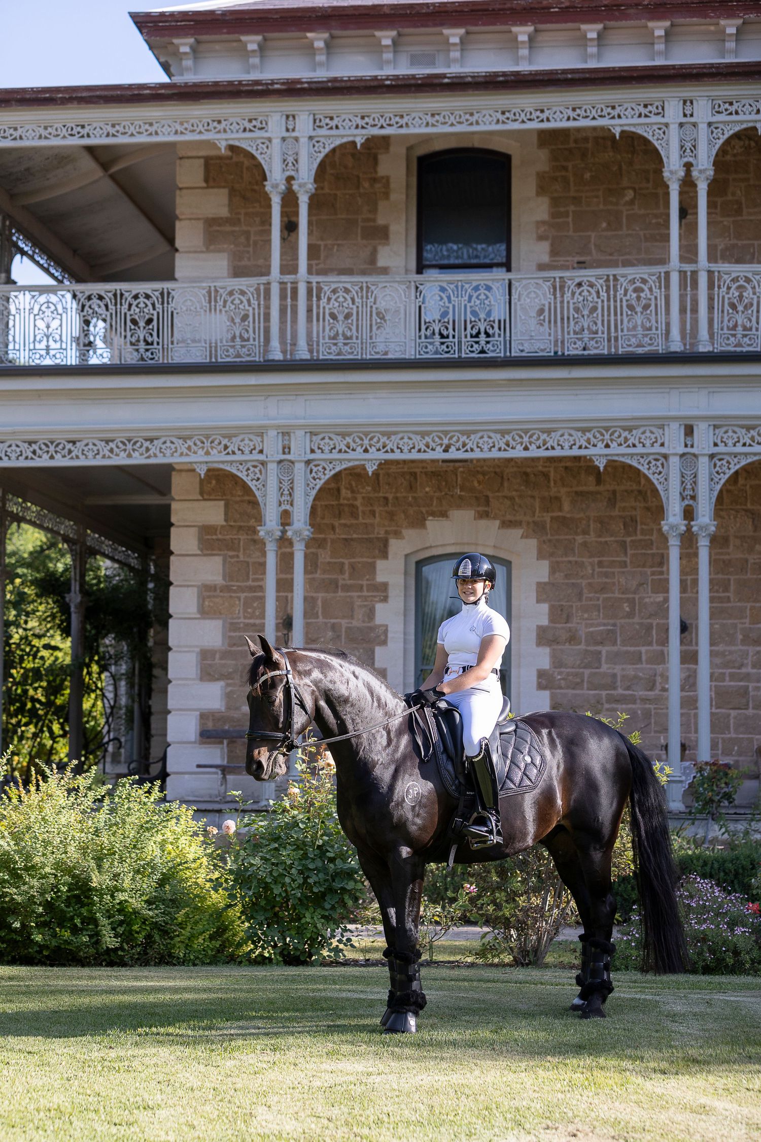 Equestrians guide their horses along the grounds of a Victorian-era mansion with decorative ironwork.