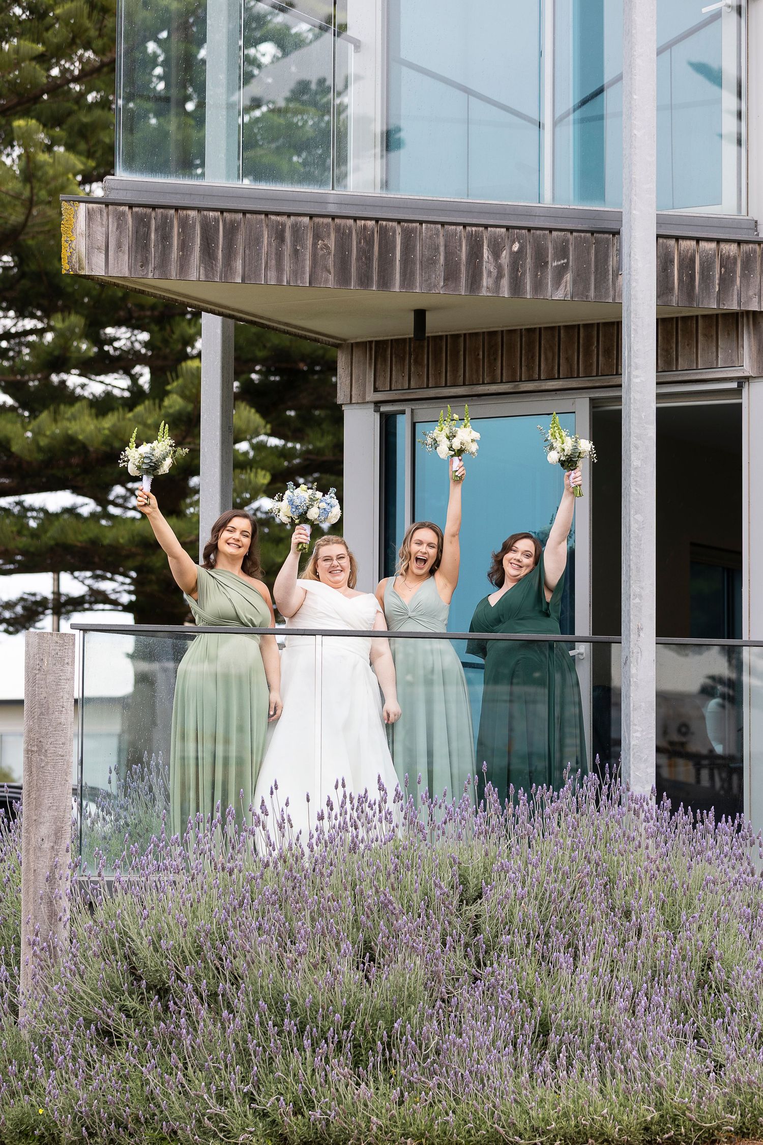 Bridesmaids in sage green dresses celebrating joyfully in front of a modern building with lavender gardens.