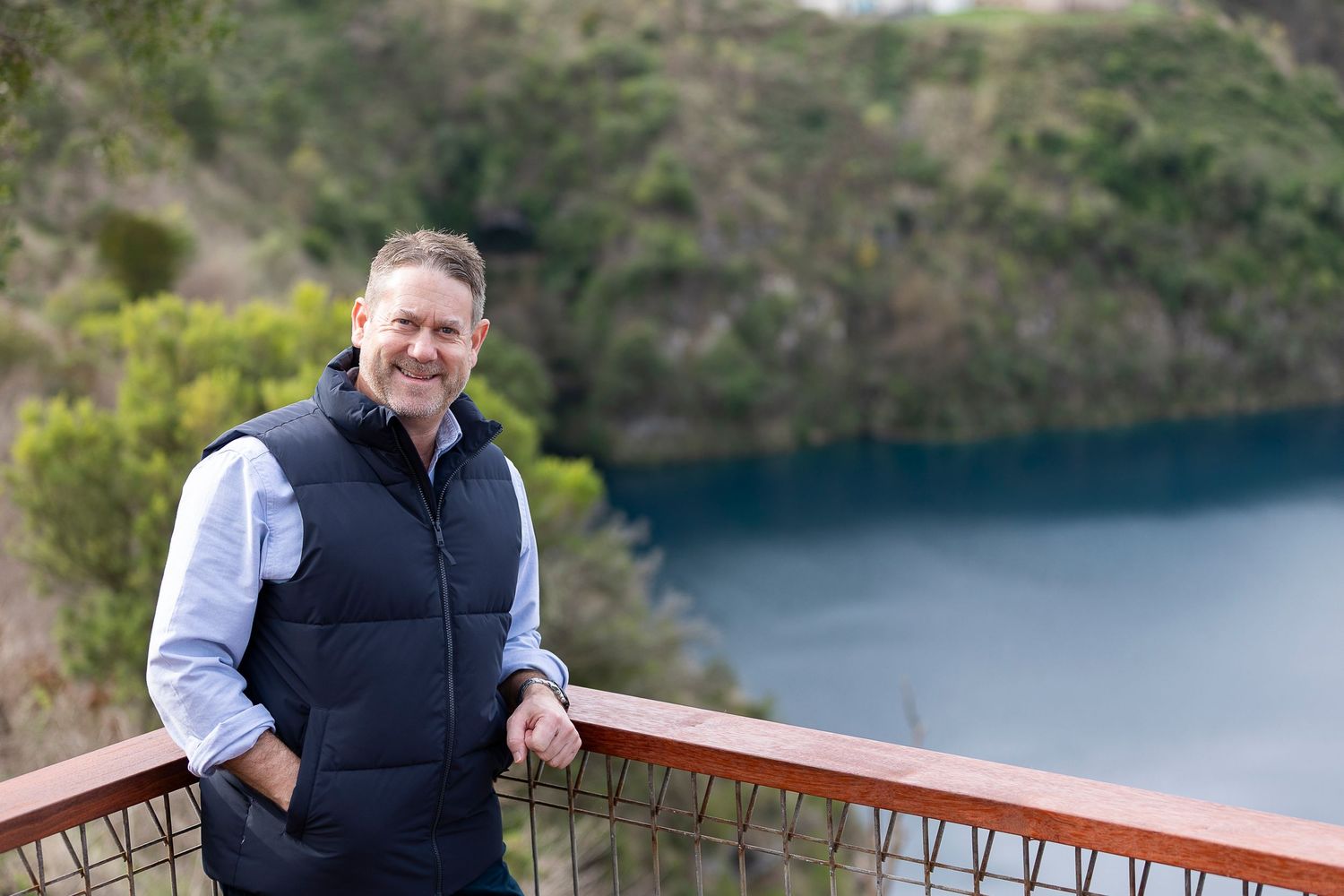 Man in navy vest smiling on wooden deck overlooking a serene blue lake surrounded by lush green hills.