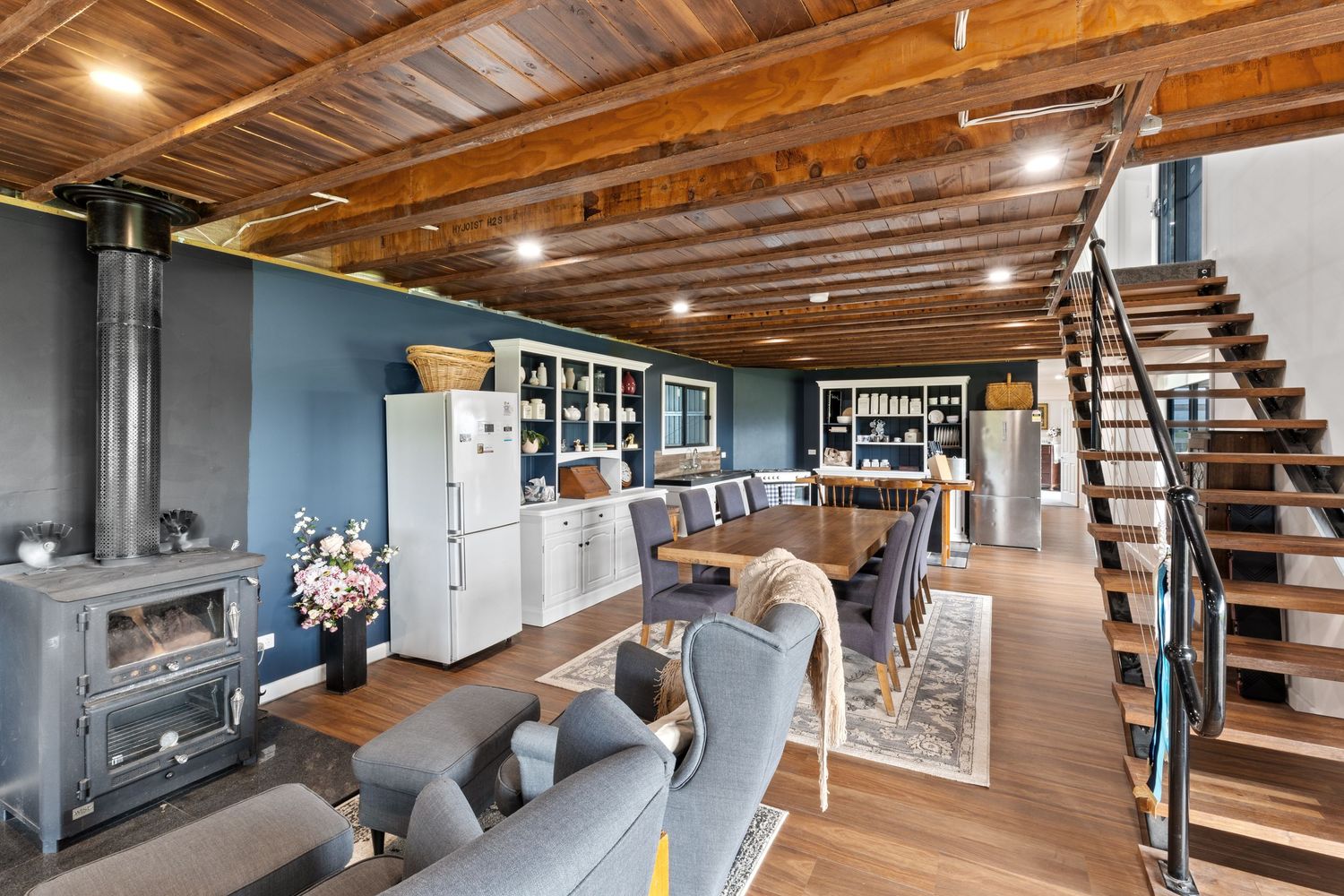 Rustic open-plan living area with wood beam ceiling, black wood stove, blue walls, dining table, and modern staircase.