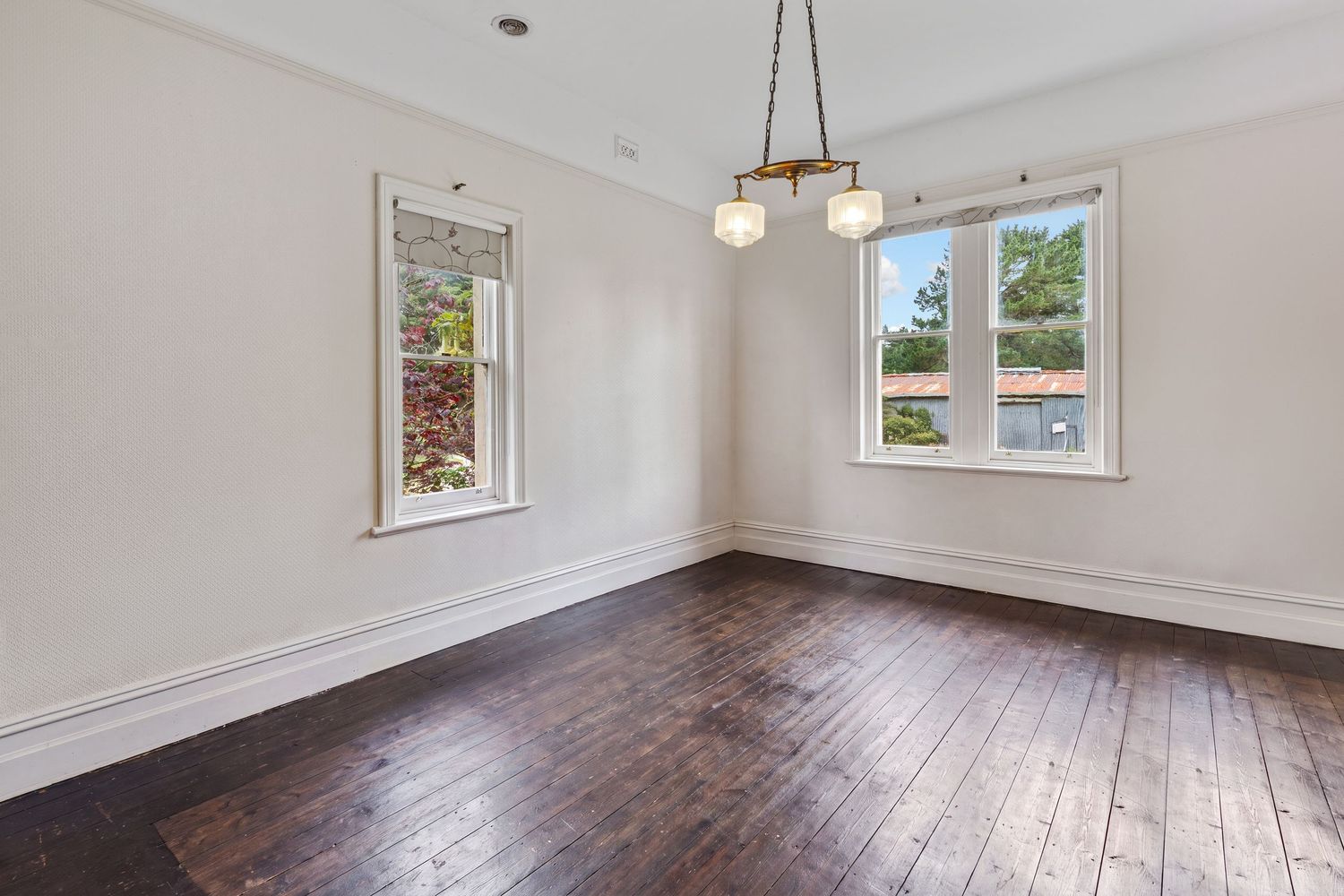 Bright empty room with dark hardwood floors, white trim, two windows, and a modern chandelier.