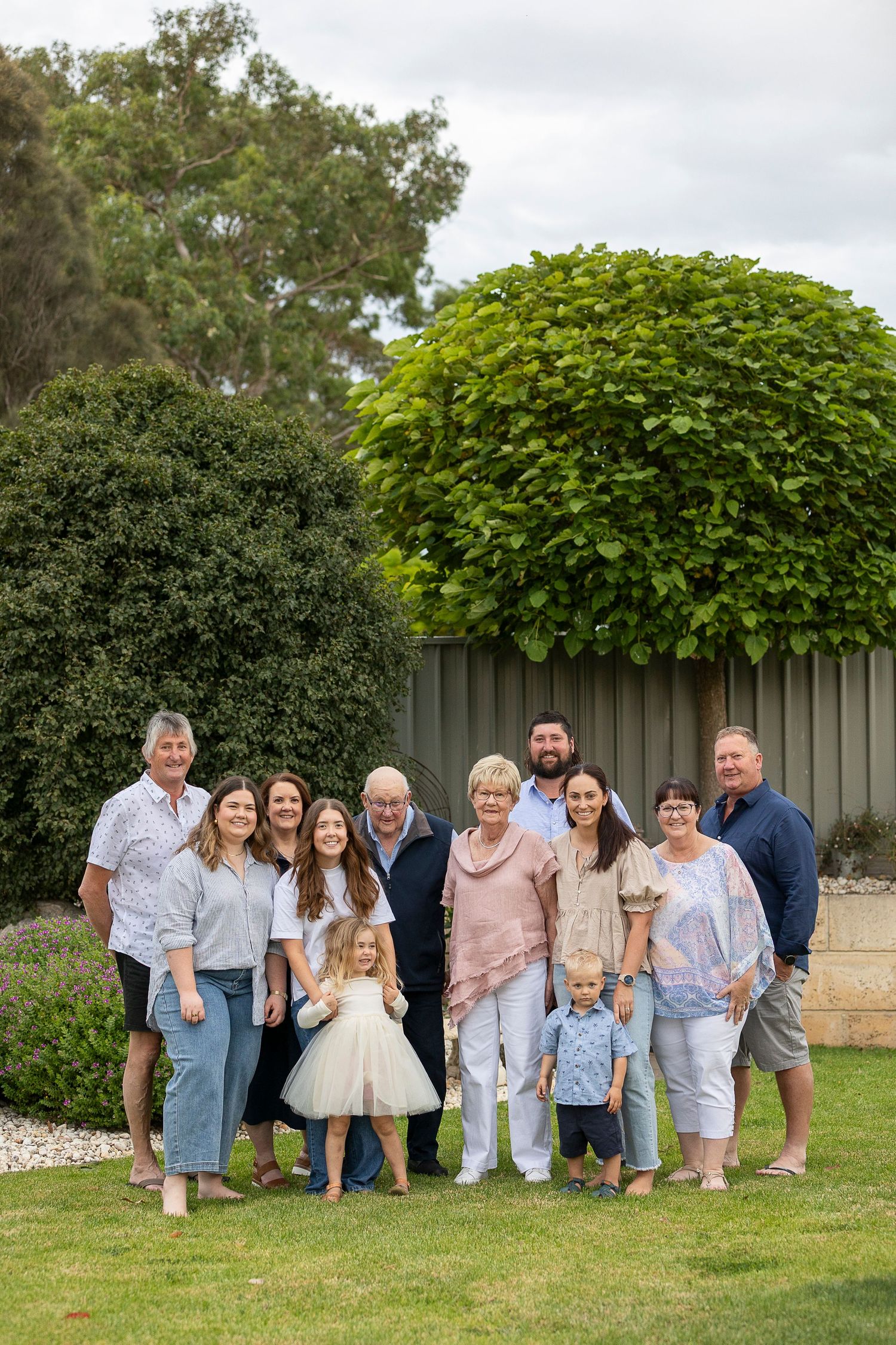 Large family group poses together in coordinated light-colored outfits against garden backdrop.