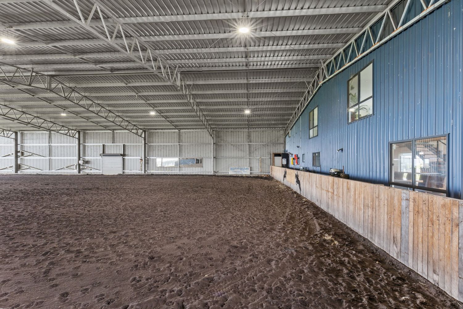 Large indoor equestrian arena with sand footing, blue steel walls, wooden kick boards, and bright overhead lighting.