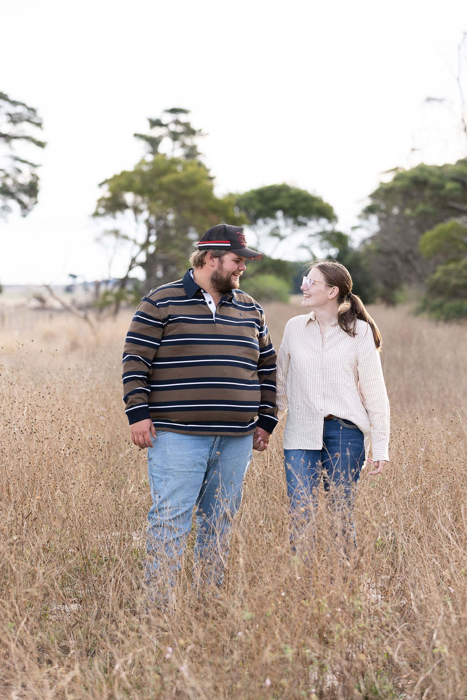 Couple walking through tall grass in a field with trees in background.