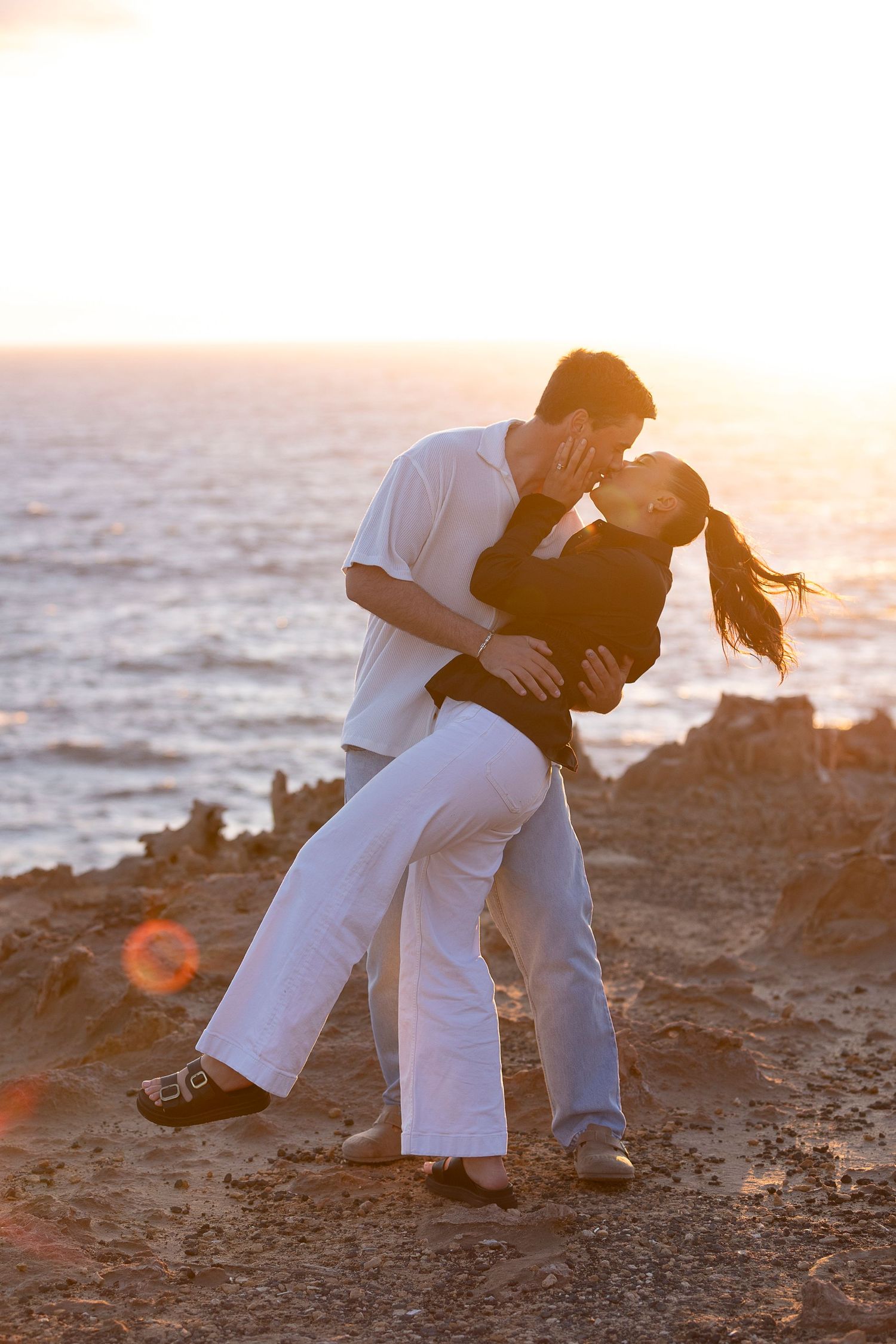 A couple in white and black outfits playfully interacts on a rocky shoreline.