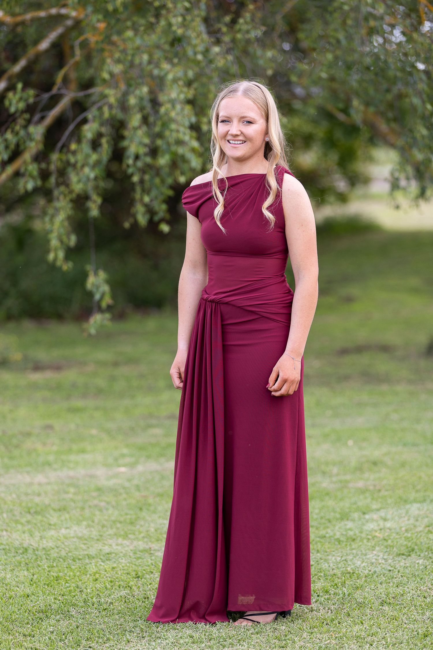 A woman in a burgundy floor-length formal dress stands on grass with trees in the background.