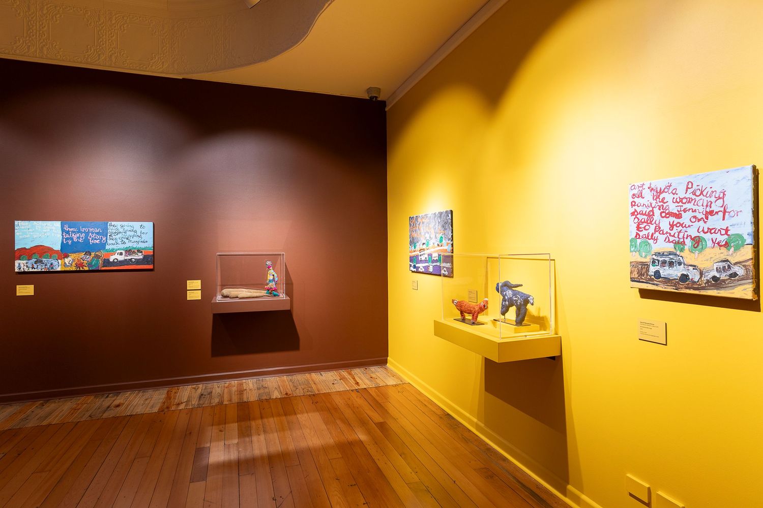 Art gallery interior with yellow walls displaying framed artwork and sculptures on red pedestals.