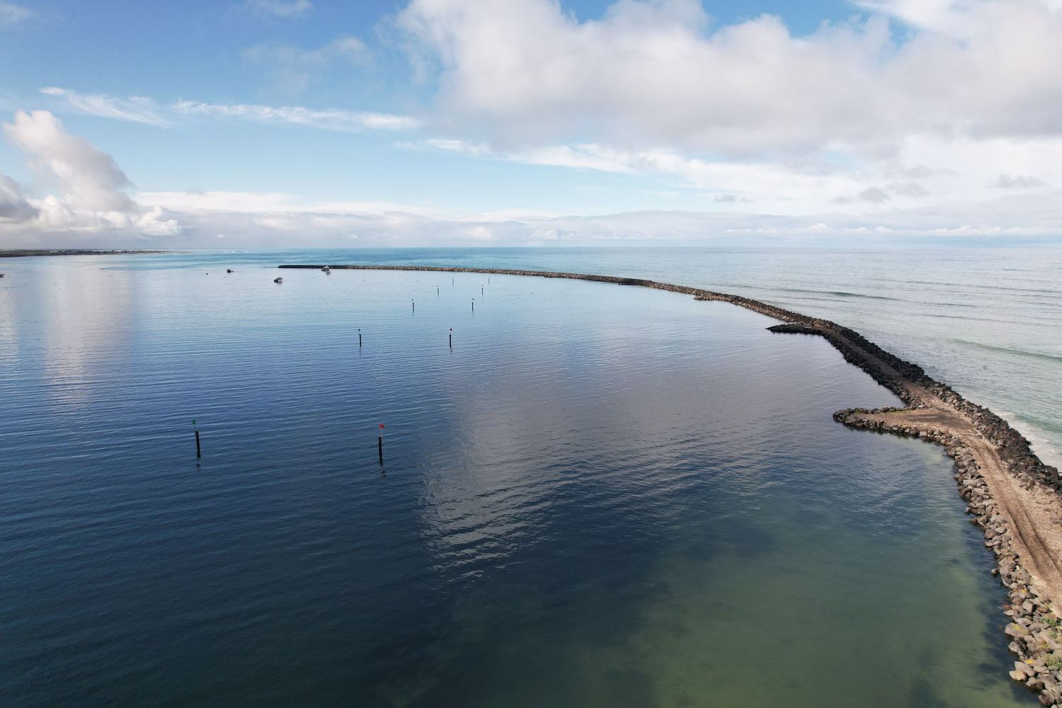 Panoramic aerial view of a curved breakwater extending into calm blue ocean waters under scattered clouds.