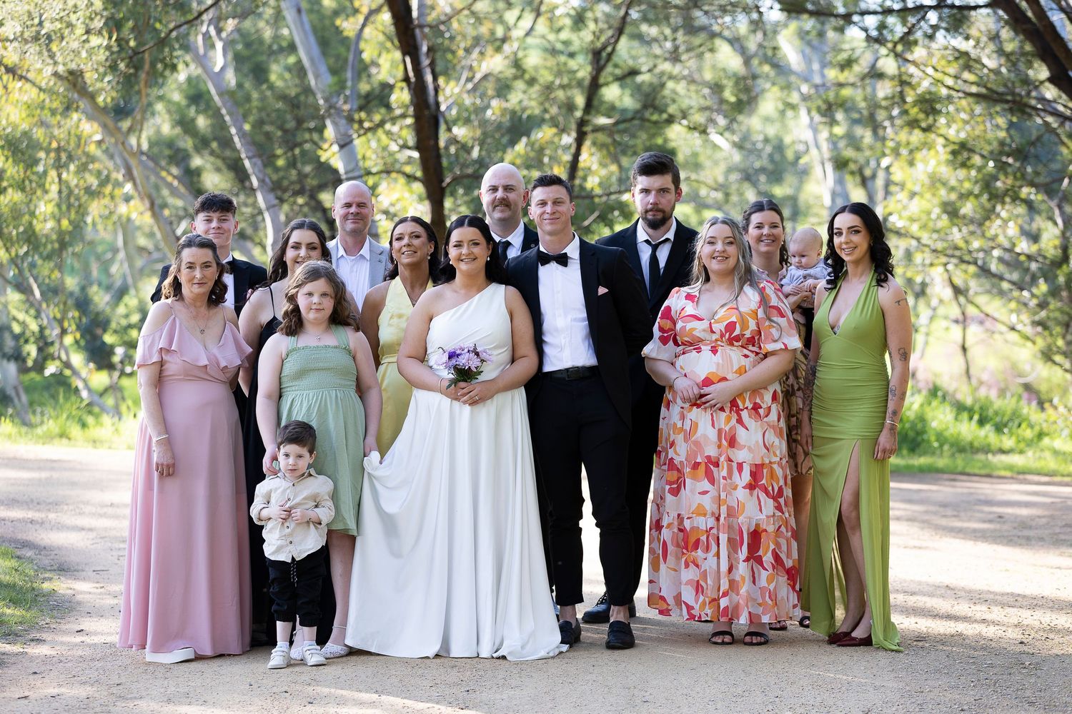Wedding party in pastel dresses poses together on a tree-lined path.