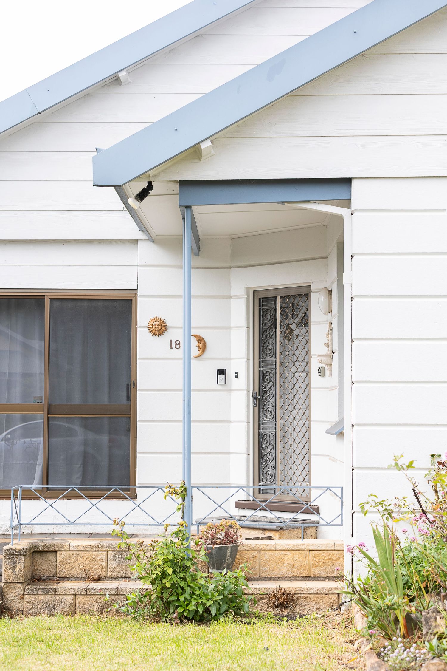 White weatherboard home exterior with blue trim, decorative screen door, stone steps, and lush garden at number 18.